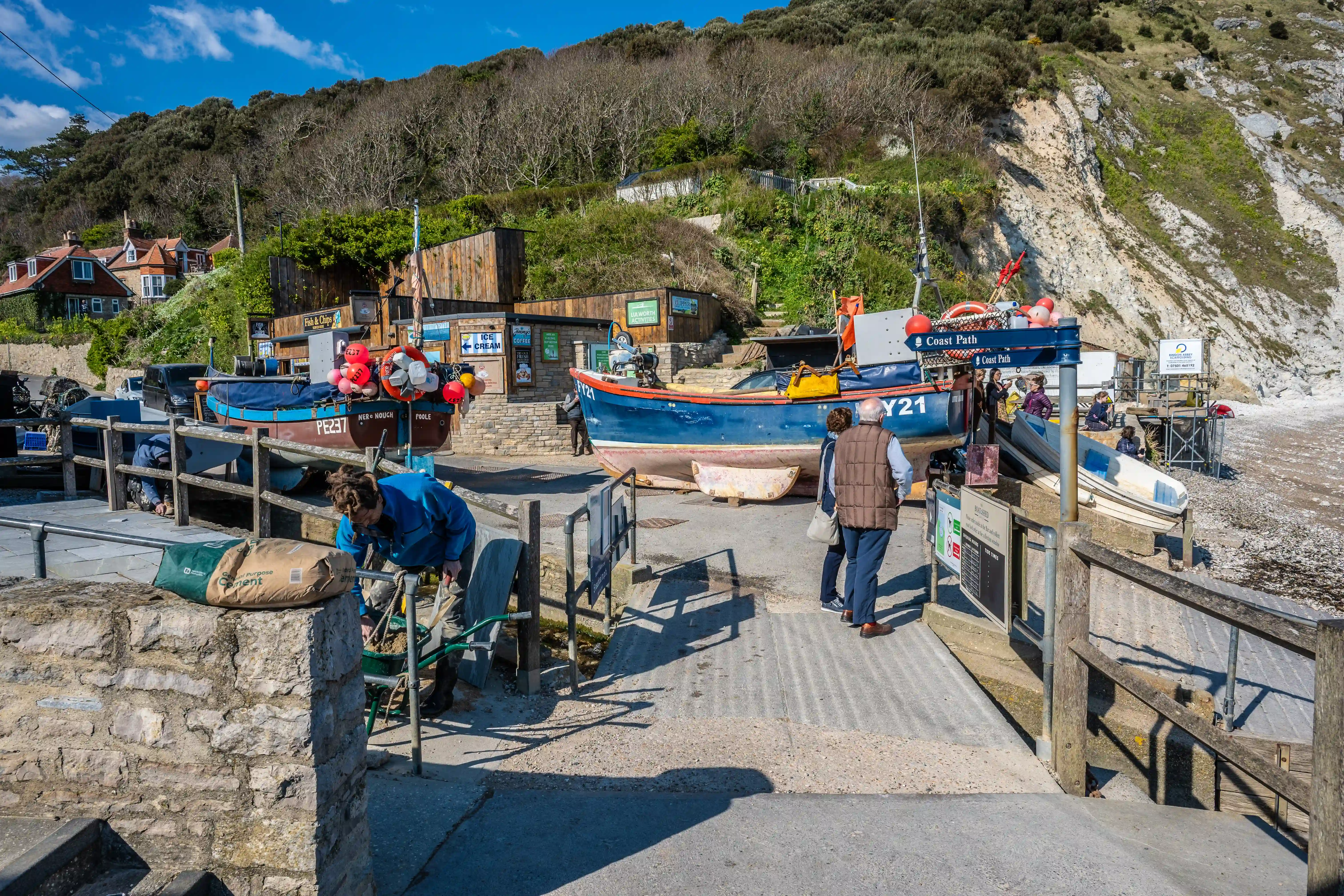 The image depicts a coastal scene with a lifeboat station and a cliffside setting. There are several people present, some walking along the pier and others engaged in activities near the boats. The lifeboat is prominently displayed with various life rings and equipment. The area is surrounded by natural cliffs and some buildings, including what appears to be an ice cream shop and other small establishments. The overall atmosphere suggests a small, bustling coastal community.