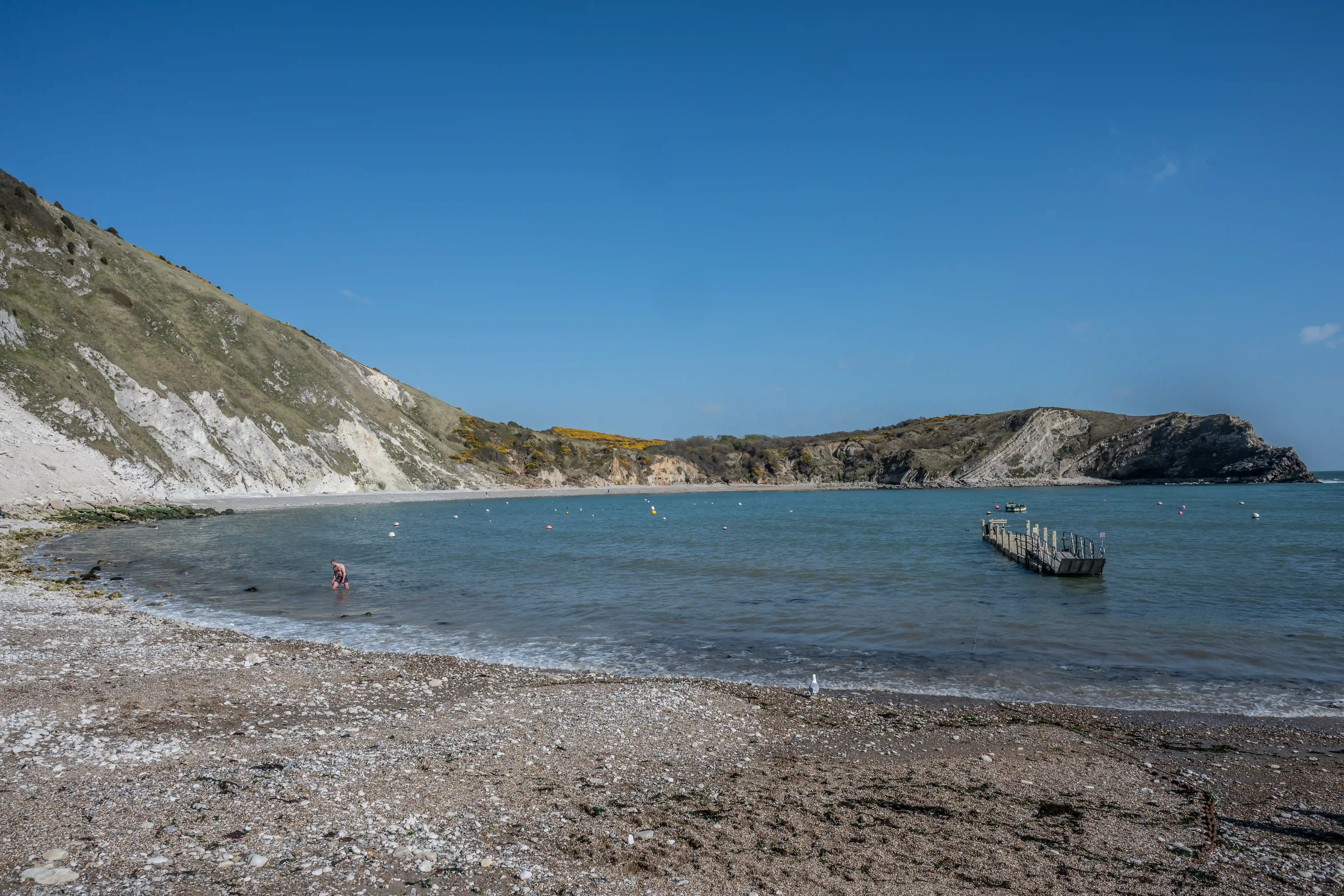 The image depicts a serene beach scene with a pebbly shore, clear blue water, and steep cliffs covered in patches of grass and shrubs. The sky is clear, and there is a wooden pier extending into the water. A person is seen wading in the shallow water near the shore, and there are buoys and a small boat visible in the distance.