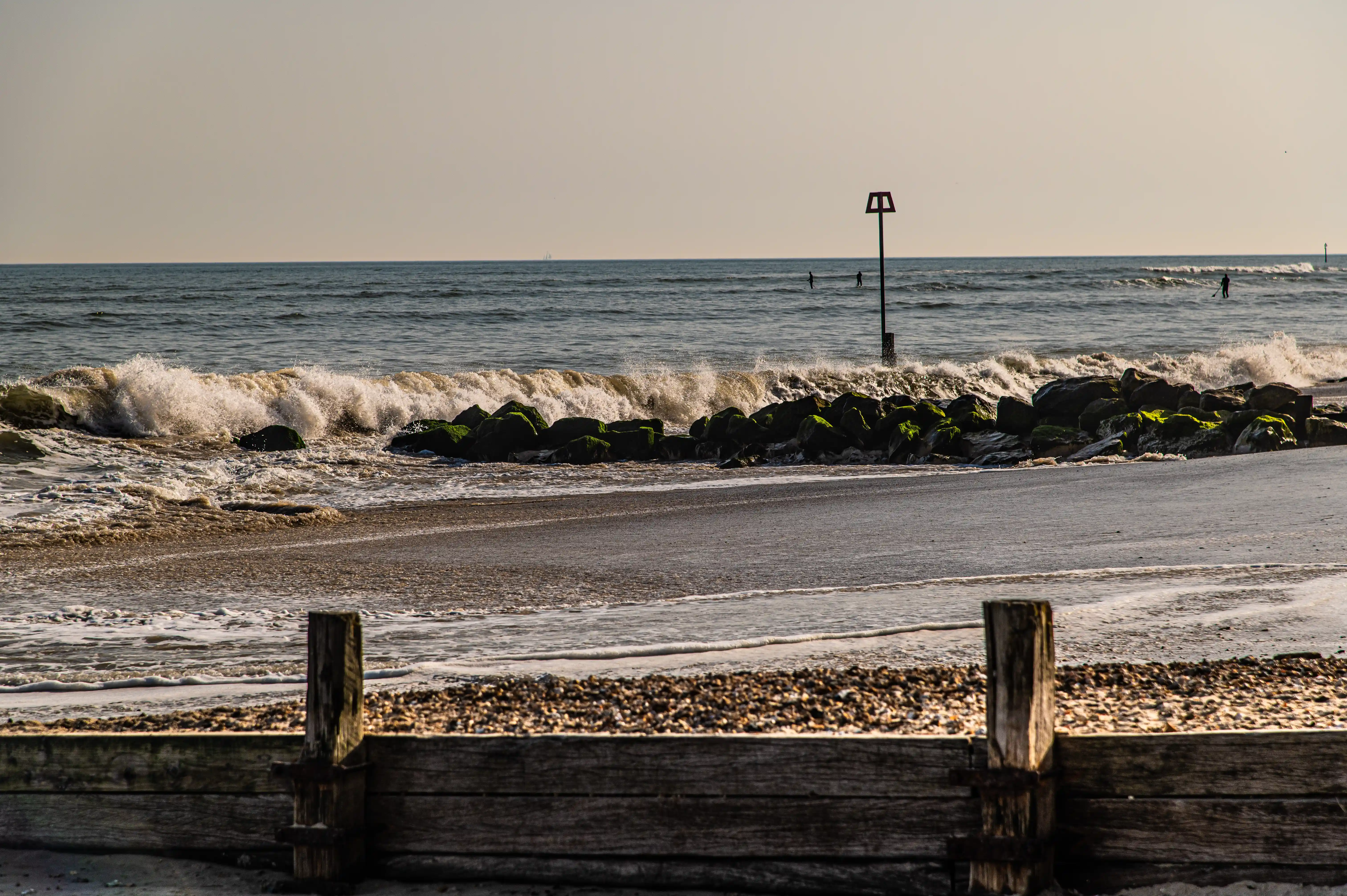 Talk about some dramatic coastal action! This shot focuses on the powerful force of the sea as waves are crashing over a line of mossy, green rocks that act as groynes. The water is churning and frothy, creating a dynamic and energetic scene. In the background, you can faintly make out some surfers catching a wave or two. The foreground features a bit of wooden barrier and pebbly shore, adding a rustic touch to the wilder ocean display.