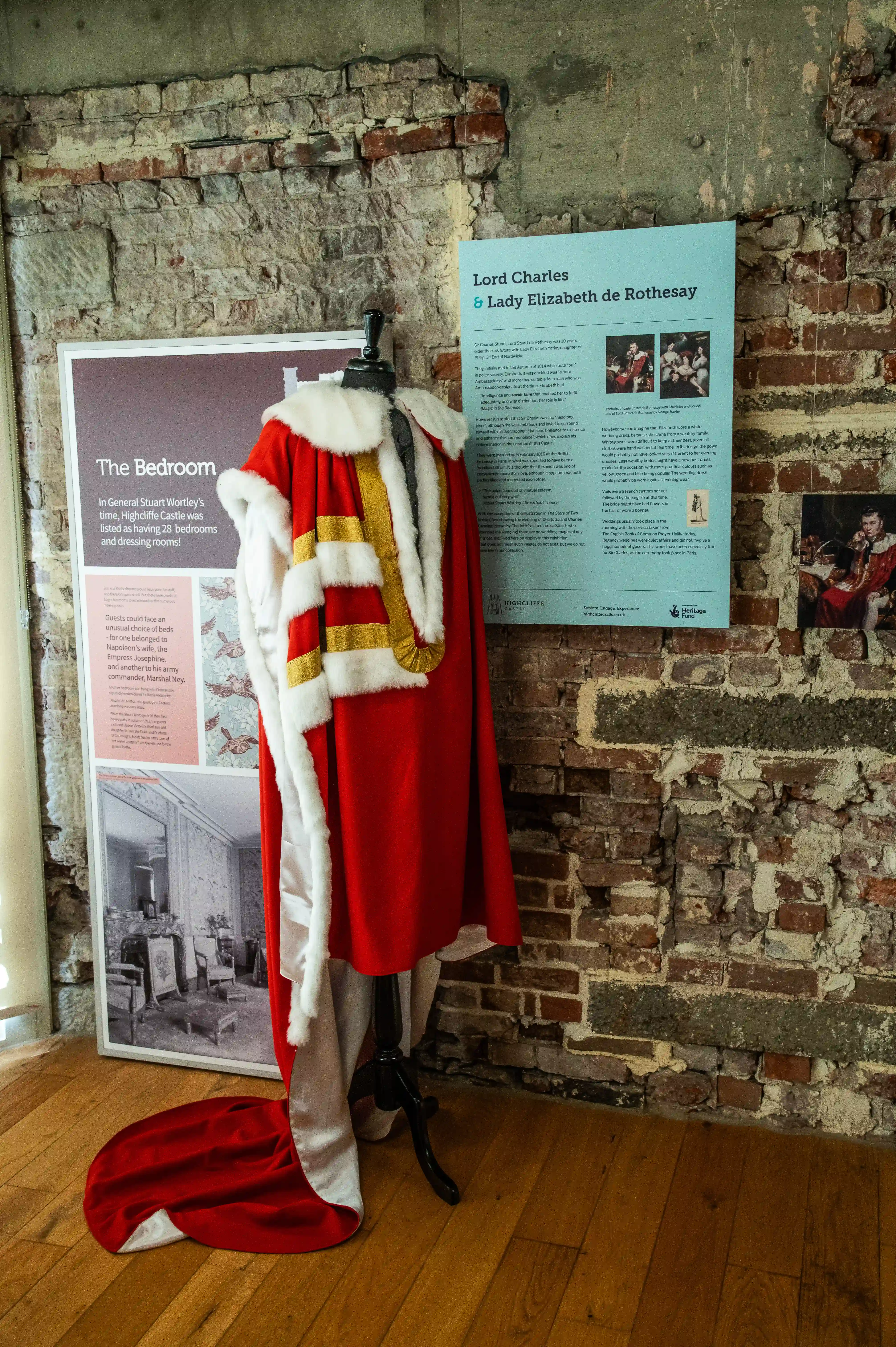 This image displays a vibrant red ceremonial robe with white fur trim and gold detailing, draped over a mannequin. It is positioned in front of a brick wall, suggesting an exhibition setting. To the left, a poster titled The Bedroom is visible, featuring an illustration of a luxurious bedroom and text describing its historical context. To the right, a light blue informational poster is present, detailing the lives of Lord Charles & Lady Elizabeth de Rothesay and including small portraits. The overall presentation hints at a historical exhibit related to royalty and their living quarters.