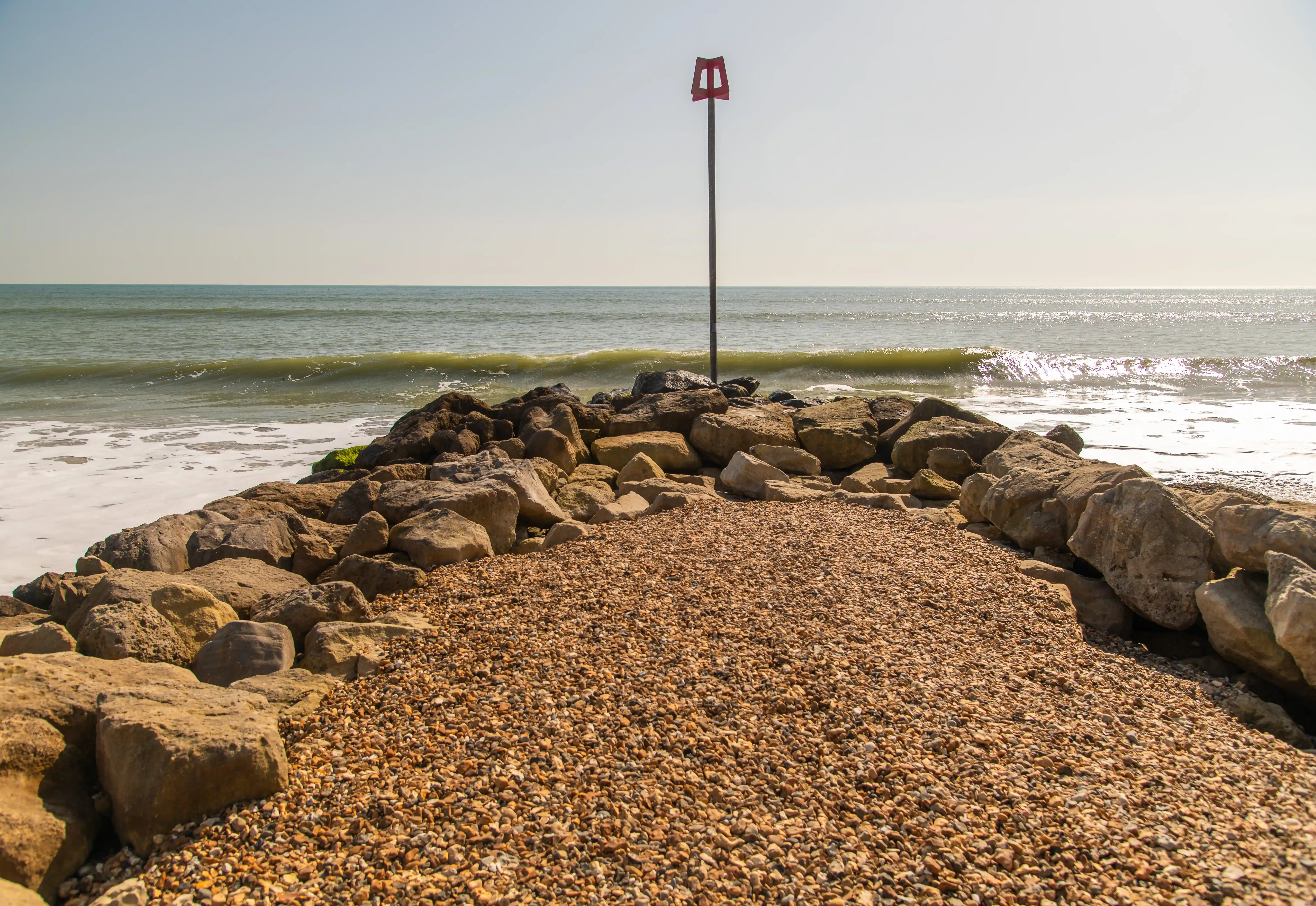 The image depicts a rocky beach with a pebbled foreground and a red marker post. The waves are gently crashing against the rocks, and the horizon is visible in the background under a clear sky.