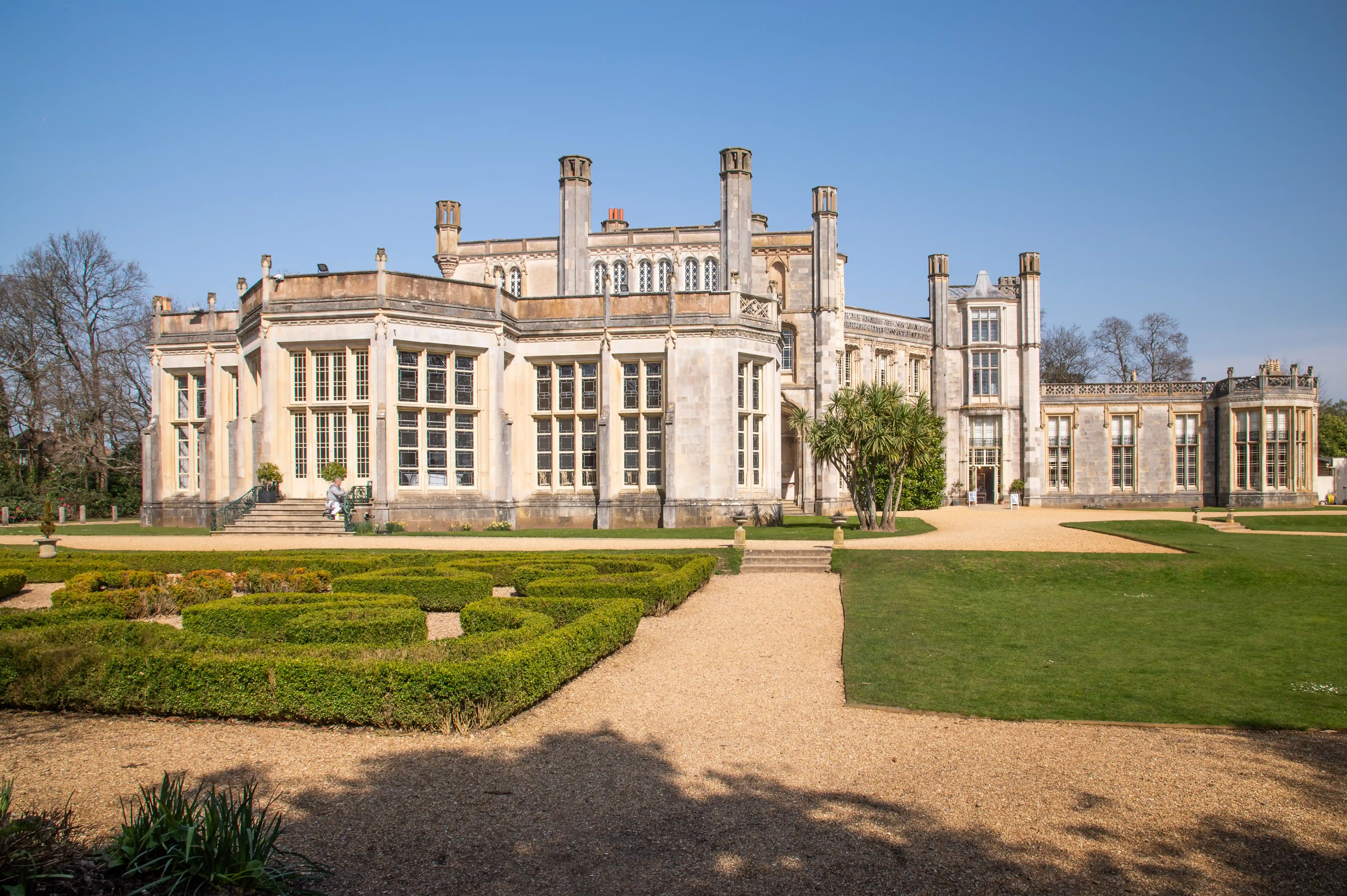 The image depicts a grand, historic building with classical architectural features, including large windows, columns, and chimneys. The structure is made of stone and has a symmetrical design. The building is surrounded by a well-maintained garden with neatly trimmed hedges, a gravel pathway, and green lawns. The sky is clear and blue, indicating a sunny day. The overall scene suggests a stately home or a significant historical building, likely located in a park or estate.
