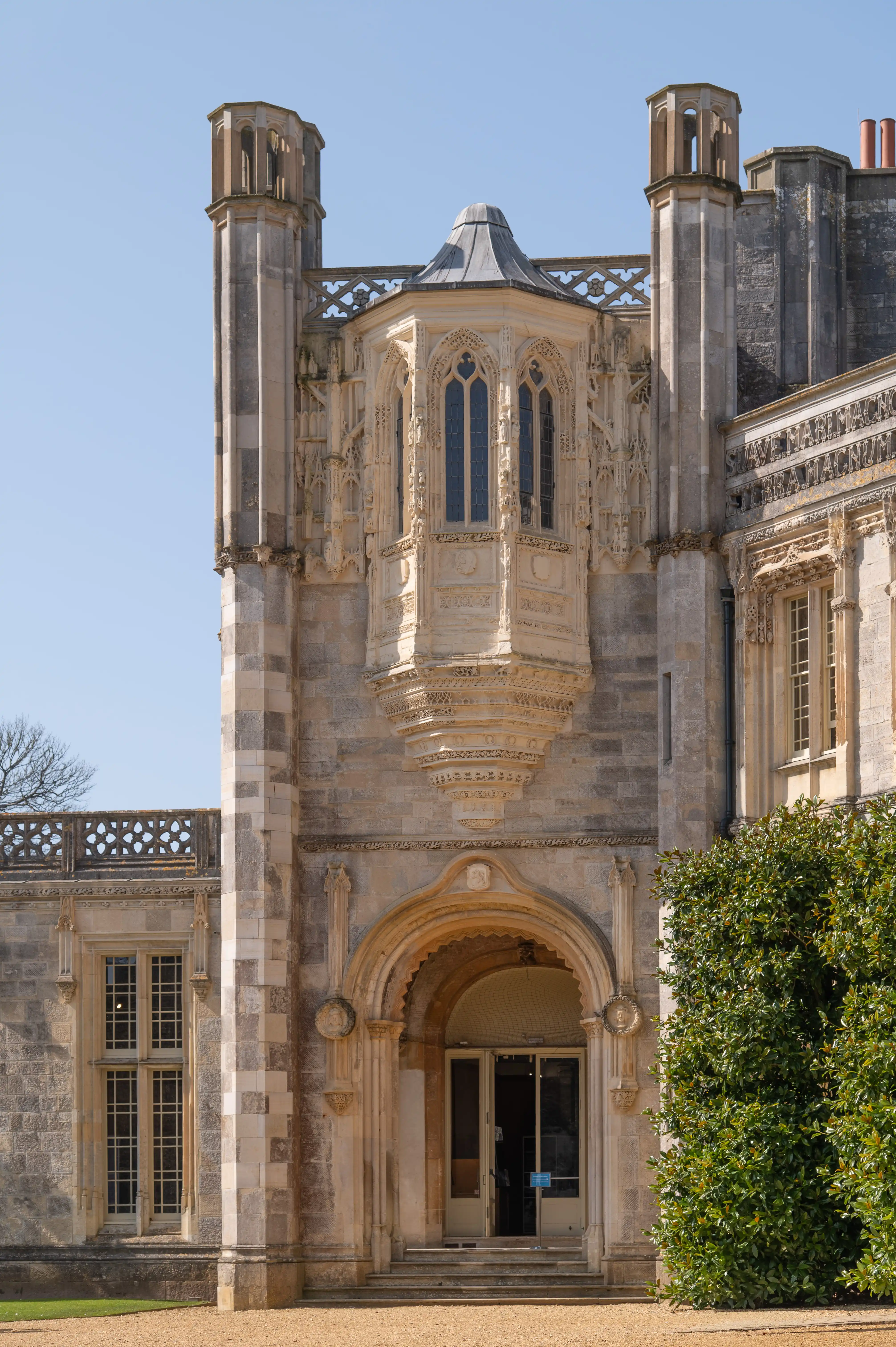 The image depicts an ornate, historical building with intricate architectural details, including tall columns, arched windows, and decorative stonework. The structure features a central entrance with a large archway and a balcony above it, flanked by two tall, rectangular towers. The building appears to be part of a larger estate or mansion, with well-maintained greenery and a clear sky in the background.
