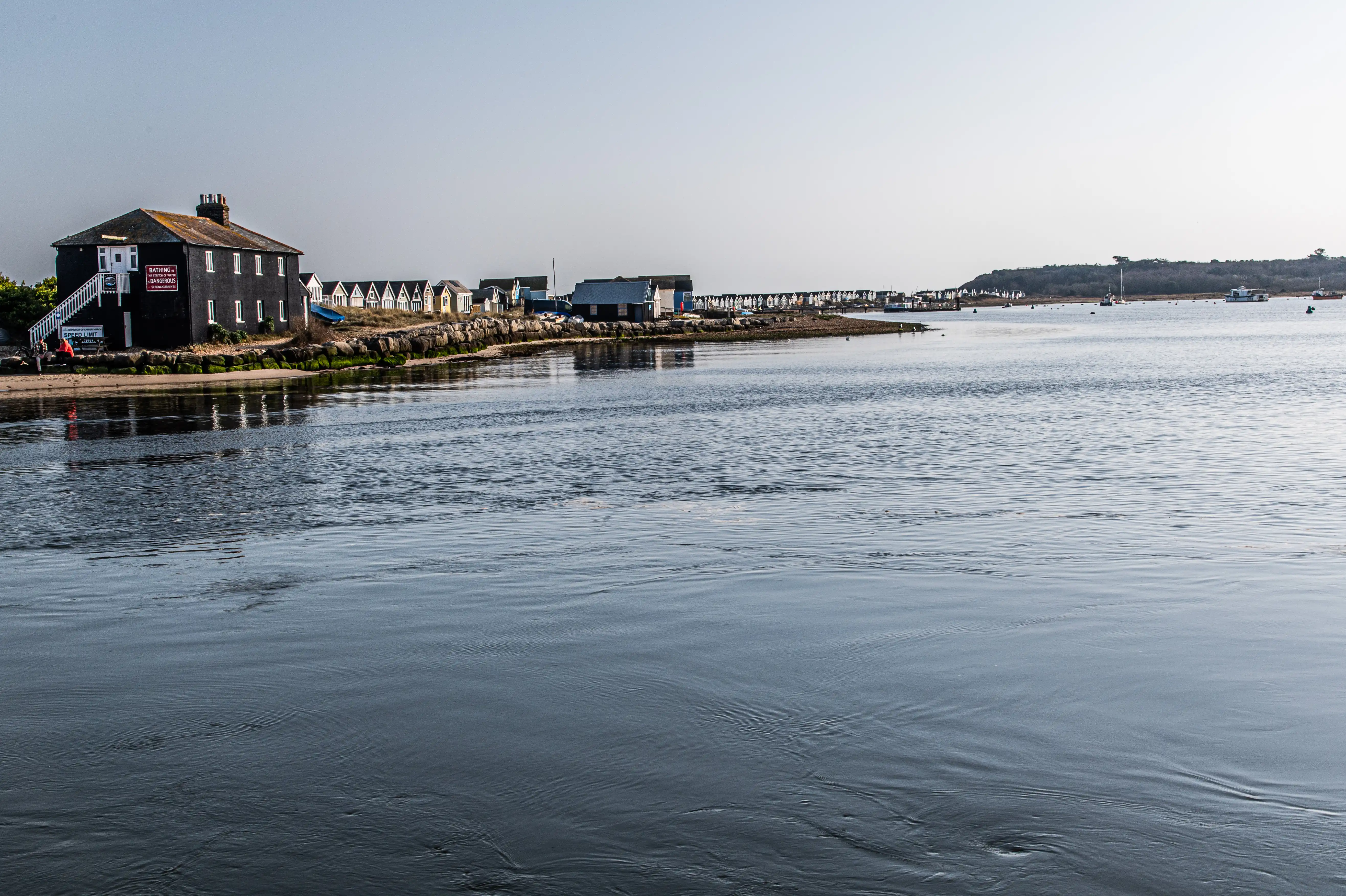 This picture captures a serene coastal scene. A dark-colored building with white trim stands prominently on the left, with stairs leading up to its entrance. Behind it, a row of charming, colorful beach huts stretches along the shoreline. The water in the foreground is calm with gentle ripples, reflecting the clear, pale sky. In the distance, you can see more structures and a few boats on the water, hinting at a small coastal community.