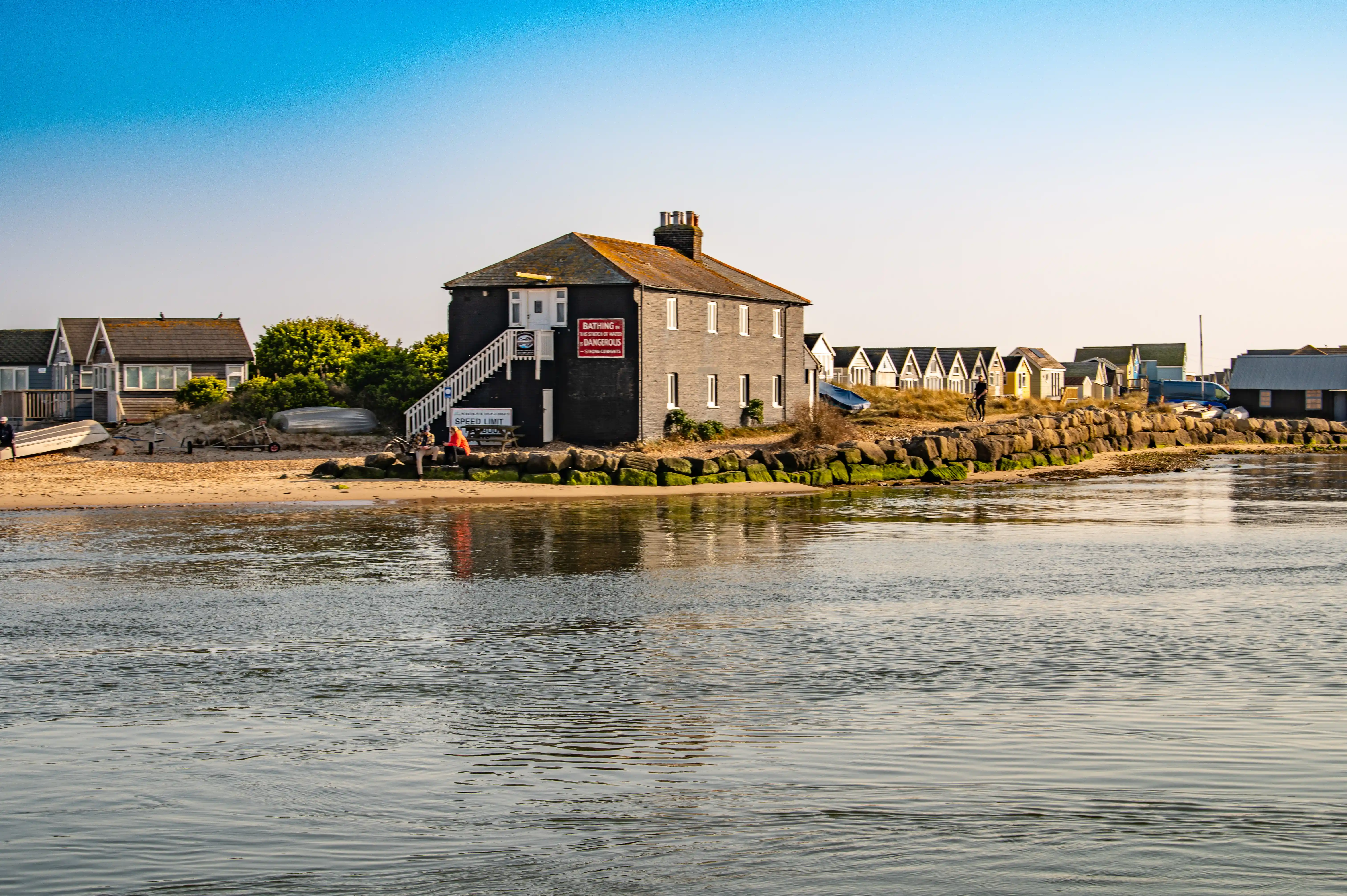 This image has a warmer, more golden light, suggesting it was taken during sunrise or sunset. The main building, similar to the one in the first image, is very clear here. The beach in front is sandy, with some boats pulled up onto it and a few people strolling around. The beach huts are also visible, lined up nicely. The water is still and reflects the soft, golden hues of the sky.