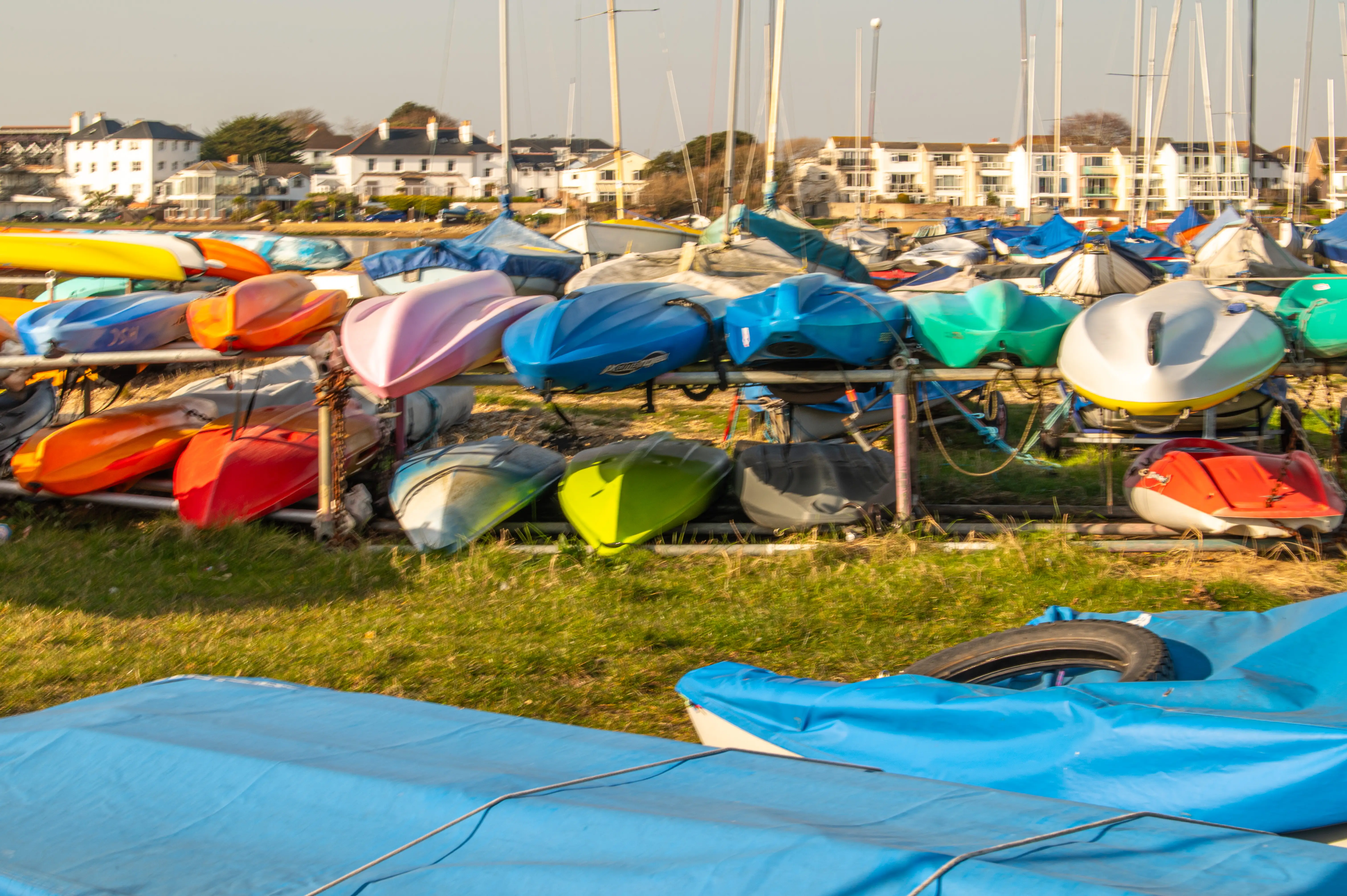 This photo captures a sunny scene of a marina or boatyard. You can see a whole collection of kayaks neatly stored on racks. They're all different bright colors – reds, oranges, blues, greens, and even some pinks and yellows – looking ready for a day on the water. In the background, there are a few buildings and a glimpse of the sea, with a few sailboat masts poking up. It gives off a relaxed, summery vibe.