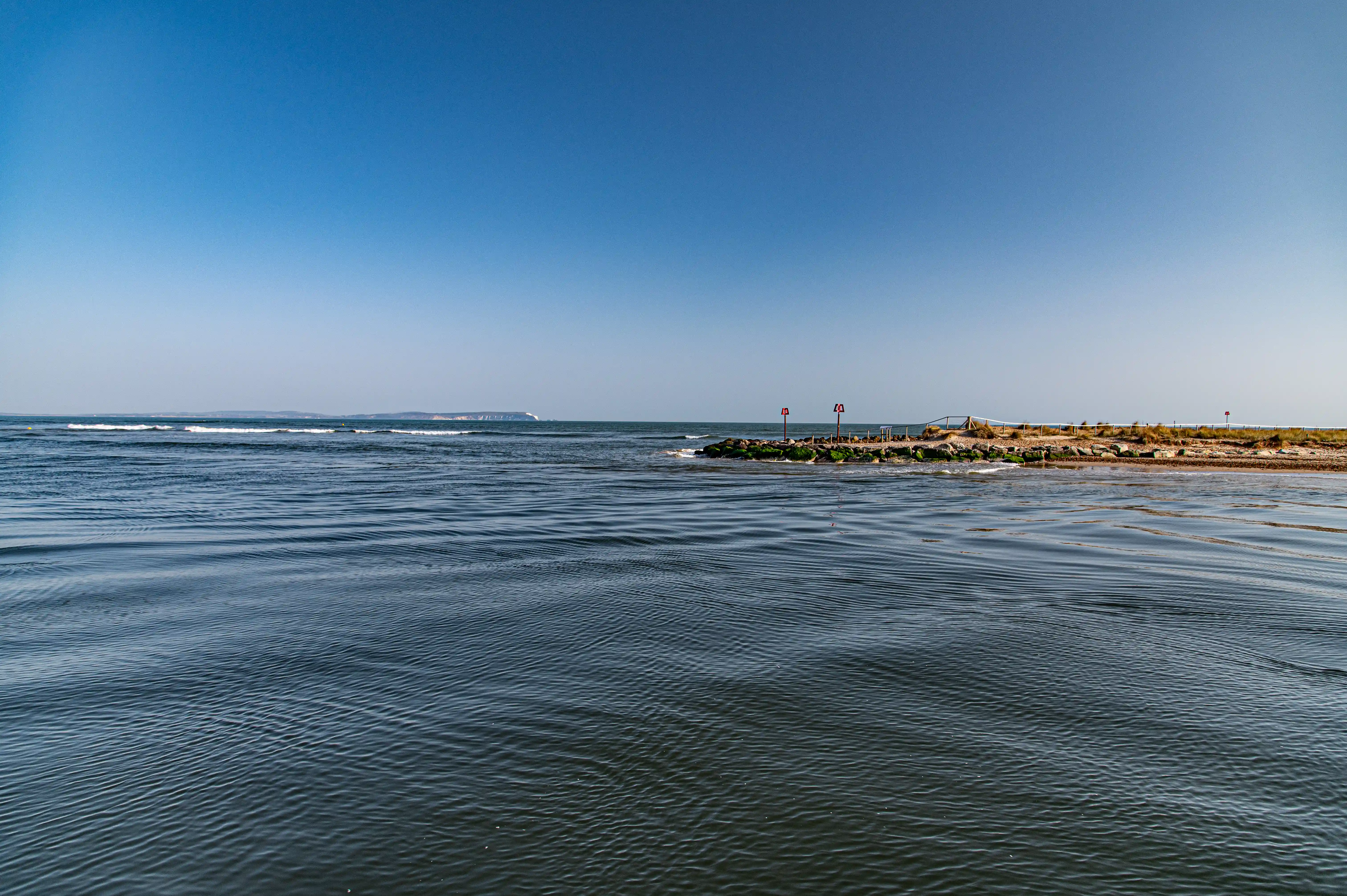 This image presents a wide, panoramic view of the sea under a clear blue sky. The water is calm with subtle wave patterns. On the right side, there's a small, rocky outcrop with some vegetation and a couple of buoys, marking a point along the coast. In the far distance, you can see a sliver of land with some white cliffs or structures, adding a sense of depth to the scene.