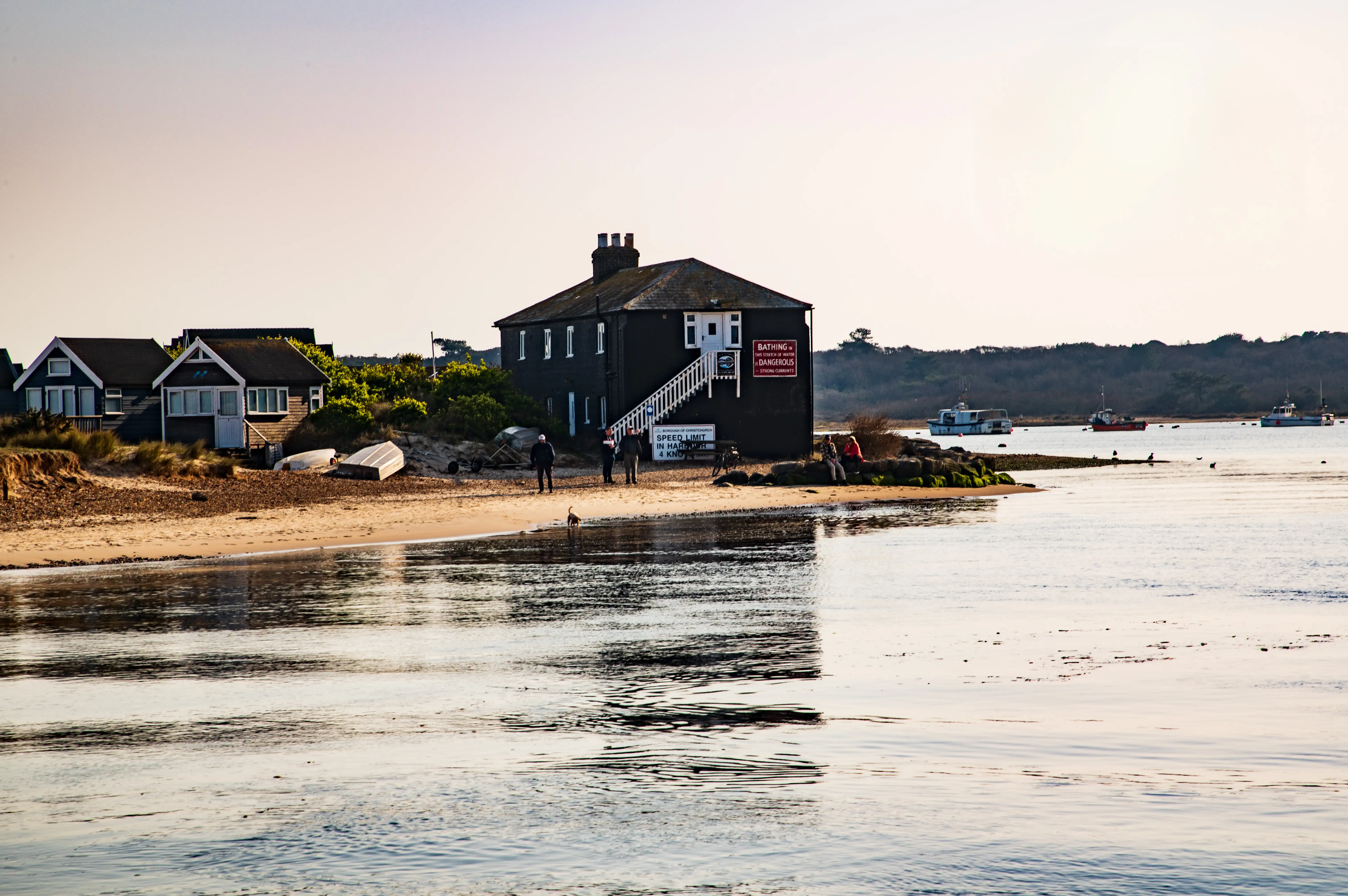 This photo has a lovely, soft light, similar to the second image, suggesting it's either early morning or late afternoon. It shows a waterfront scene with the main building and some smaller houses. The water is very calm, creating a beautiful reflection of the sky and the buildings. A few boats are moored in the water in the background. You can see some people on the beach and a few birds near the water's edge.