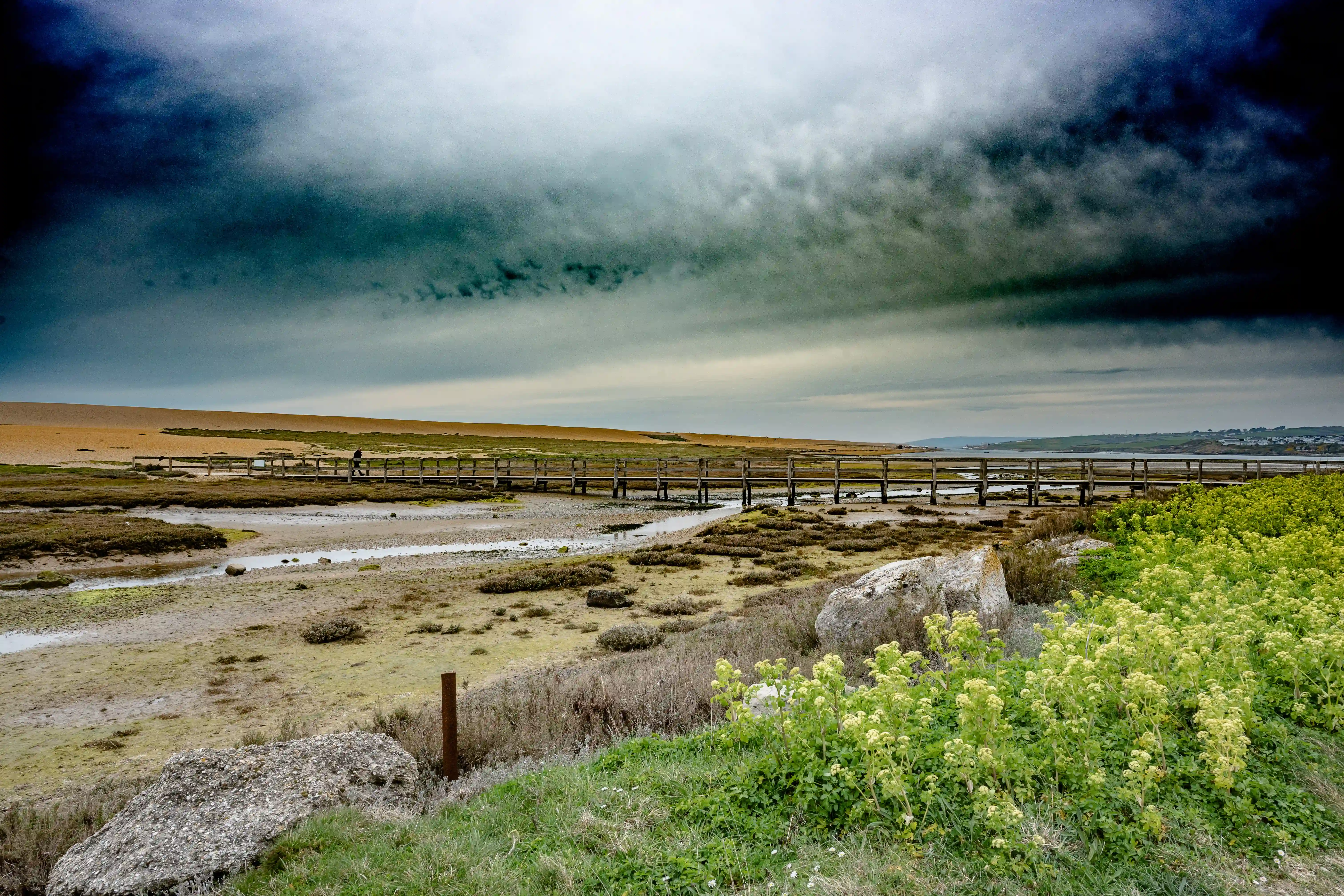 The image depicts a scenic landscape with a wooden boardwalk extending into the distance. The sky is filled with dark, ominous clouds, suggesting an impending storm. The terrain is a mix of sandy and grassy areas with patches of vegetation. In the background, there are rolling hills and a distant view of the sea. The overall atmosphere is one of tranquility and natural beauty, with a sense of solitude and serenity.