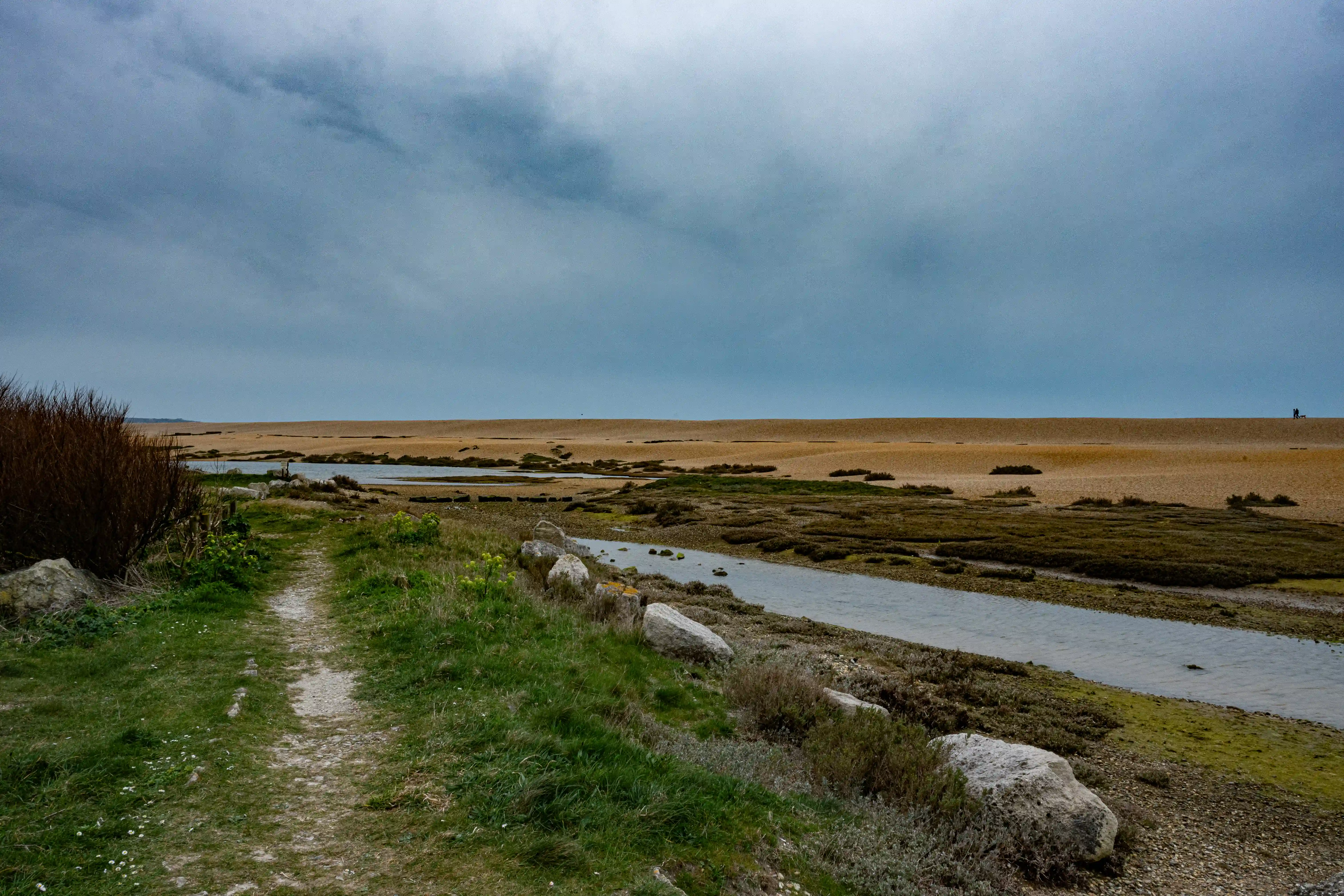 The image depicts a natural landscape featuring a narrow dirt path leading towards a vast, flat, sandy area under a cloudy sky. The path is bordered by green vegetation and small rocks. A shallow stream runs parallel to the path, reflecting the overcast sky. The scene conveys a sense of tranquility and isolation, with minimal human presence.