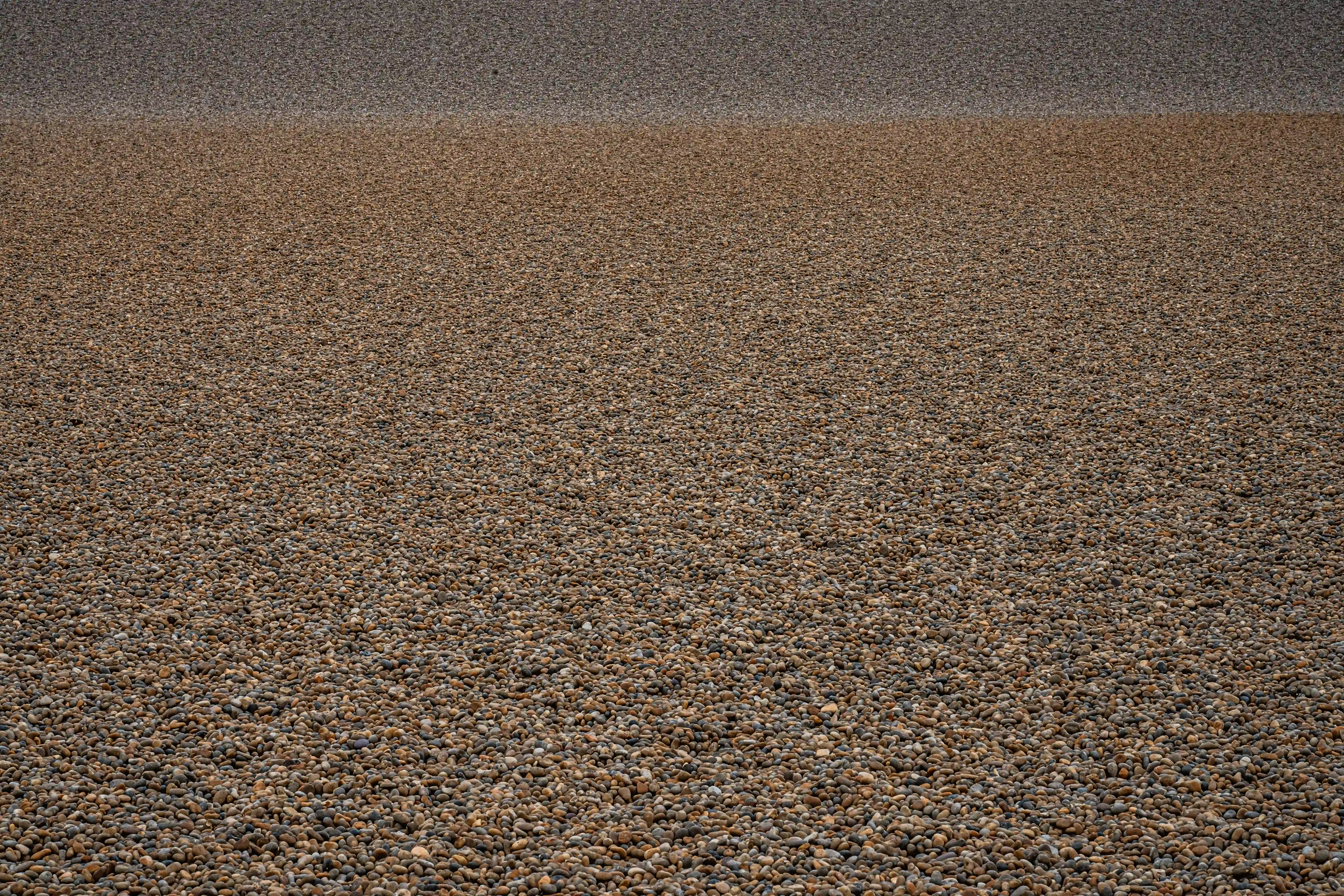 The image shows a close-up view of a surface covered with small, rounded pebbles. The pebbles are of varying shades of brown and gray, and the surface appears to be composed of multiple layers of these pebbles, with a noticeable transition between two distinct layers. The texture and color of the pebbles suggest a natural, outdoor setting, possibly a gravel path or a beach.