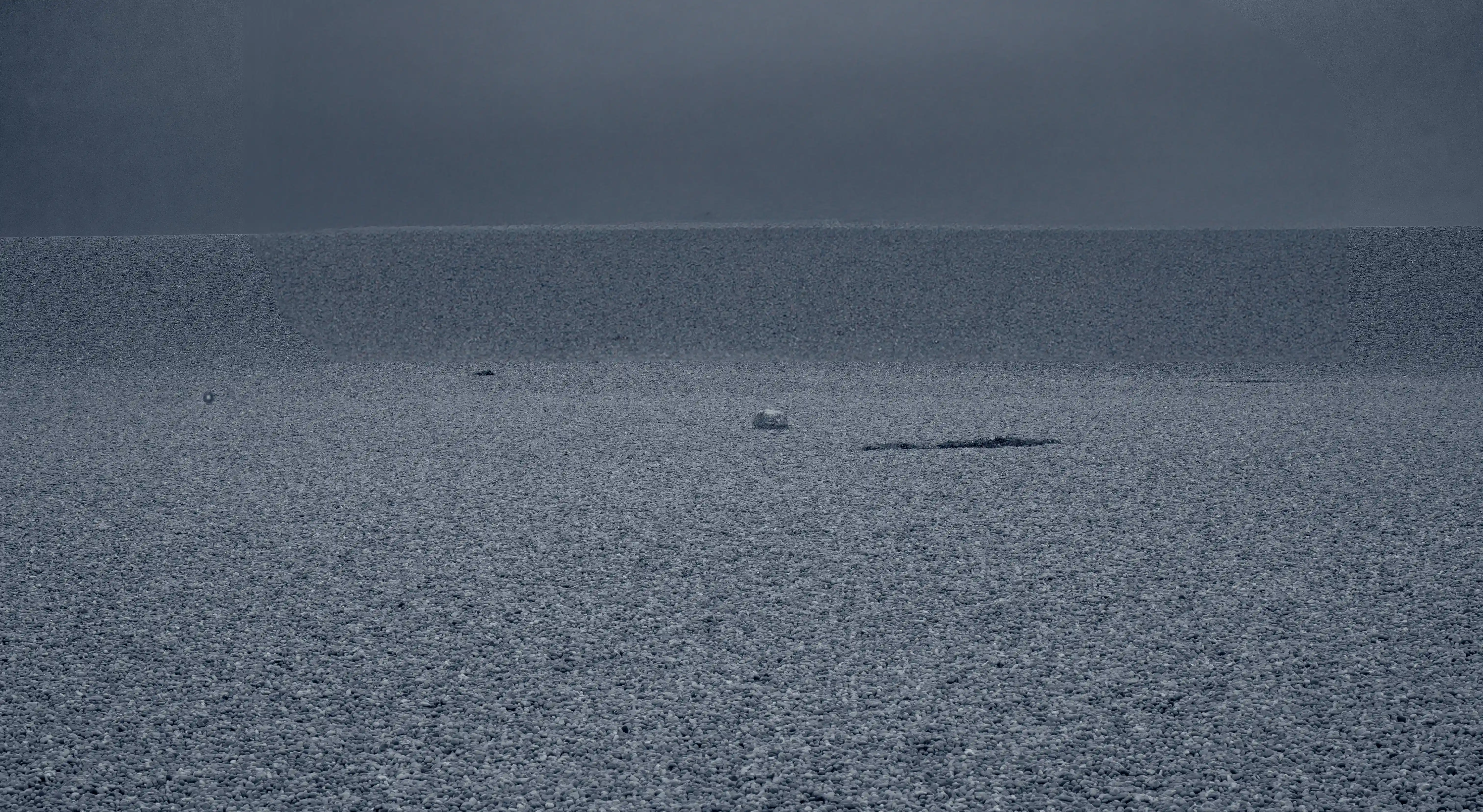 The image depicts a vast, textured landscape, a pebble beach, under a dark, overcast sky. The ground is covered with small, uniform grains or crystals, creating a rippled pattern. There are a few small objects scattered across the landscape, including what appears to be a small white object and some dark patches. The overall atmosphere is somber and desolate, with a sense of vastness and isolation.