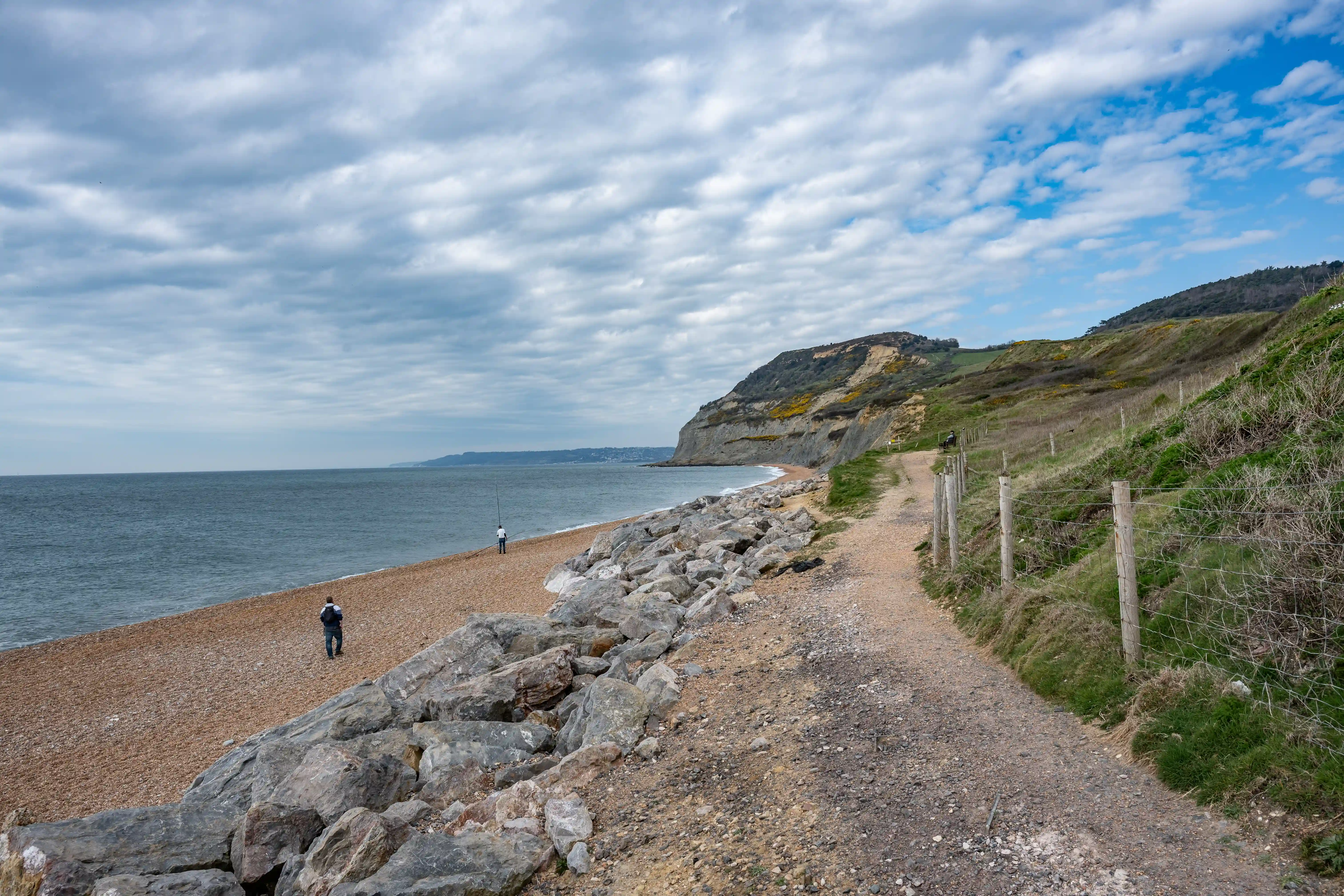 The image depicts a coastal scene with a pebble beach, a rocky path, and a grassy hillside. The sky is partly cloudy with patches of blue. Two individuals are visible: one standing on the beach and the other further along the path. The coastline extends into the distance with cliffs and a stretch of water.