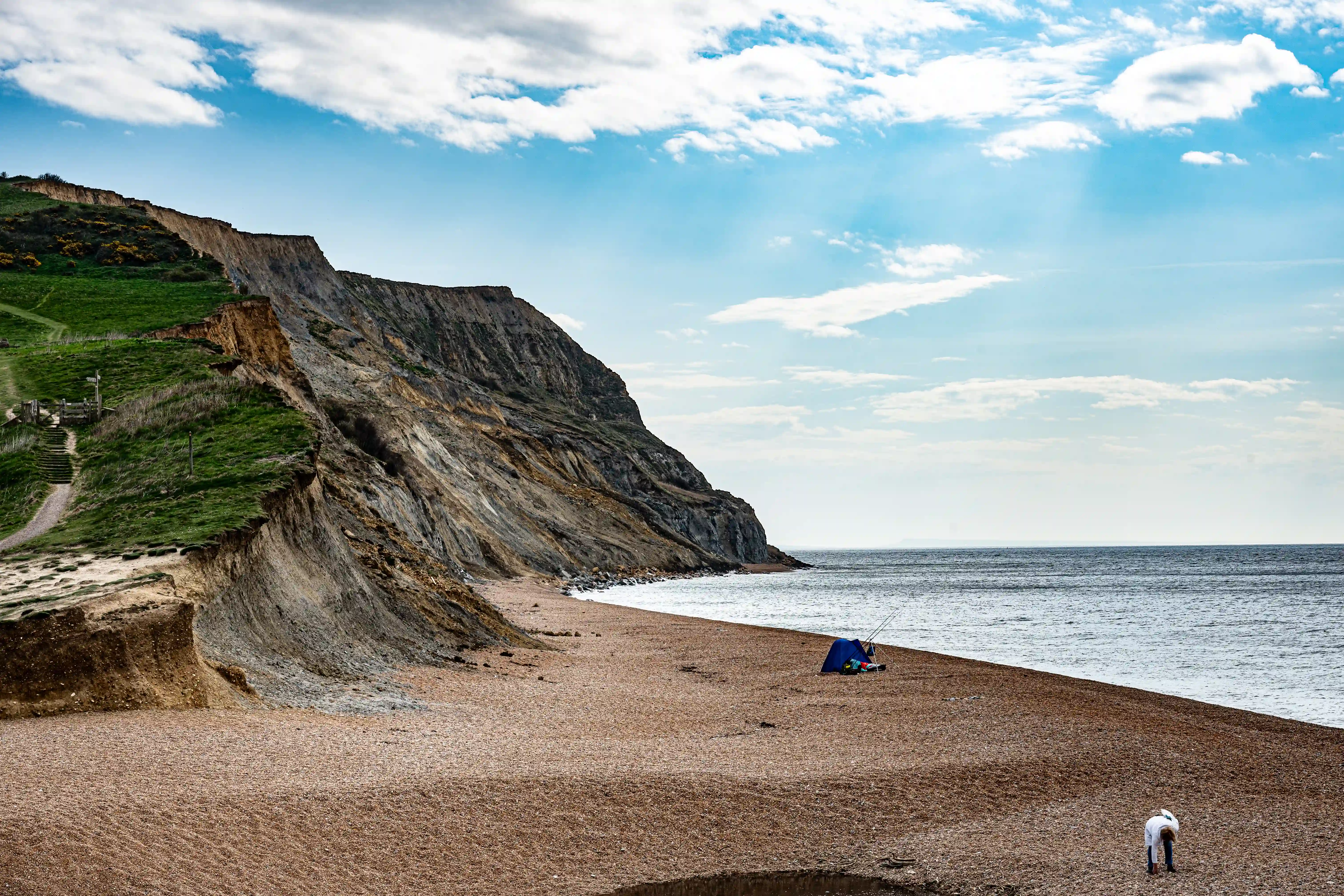 The image depicts a serene beach scene with steep, grassy cliffs on the left and a pebbly shoreline extending to the right. The sky is mostly clear with some scattered clouds. A person is seen walking along the beach, and there is a tent or shelter set up near the water's edge. The overall atmosphere is calm and peaceful, suggesting a quiet, possibly remote location ideal for relaxation or fishing.