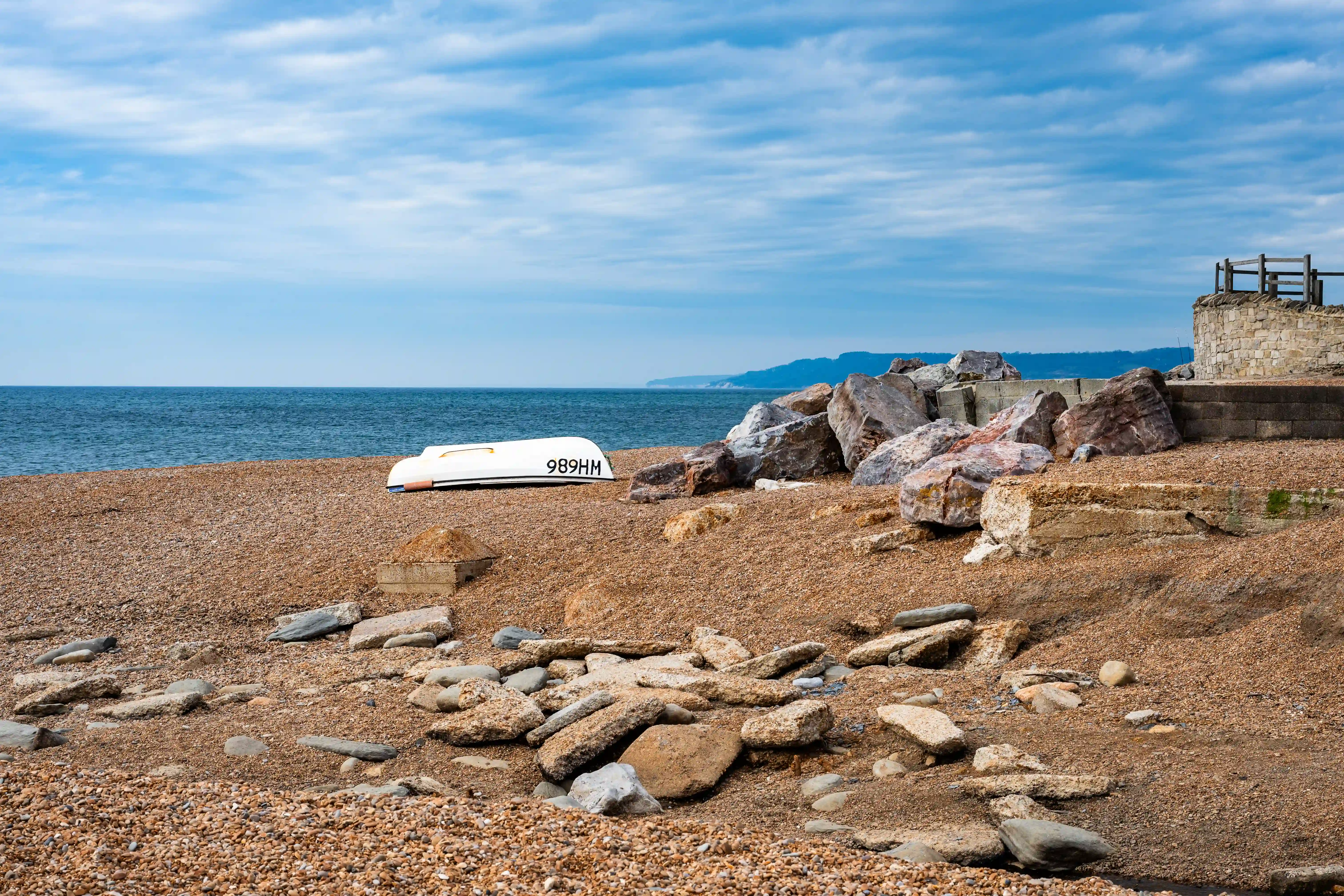 This is a bit more of a serene shot. It's a pebble beach with a white boat, which looks like it's had its registration number, '989HM', painted on the side, pulled up onto the shore. There are some substantial rocks and bits of concrete, perhaps part of an old sea defence or structure, giving it a bit of an industrial feel against the natural landscape. In the distance, you can see the hazy outline of hills along the coast. The sky is a clear blue with a few wispy clouds.