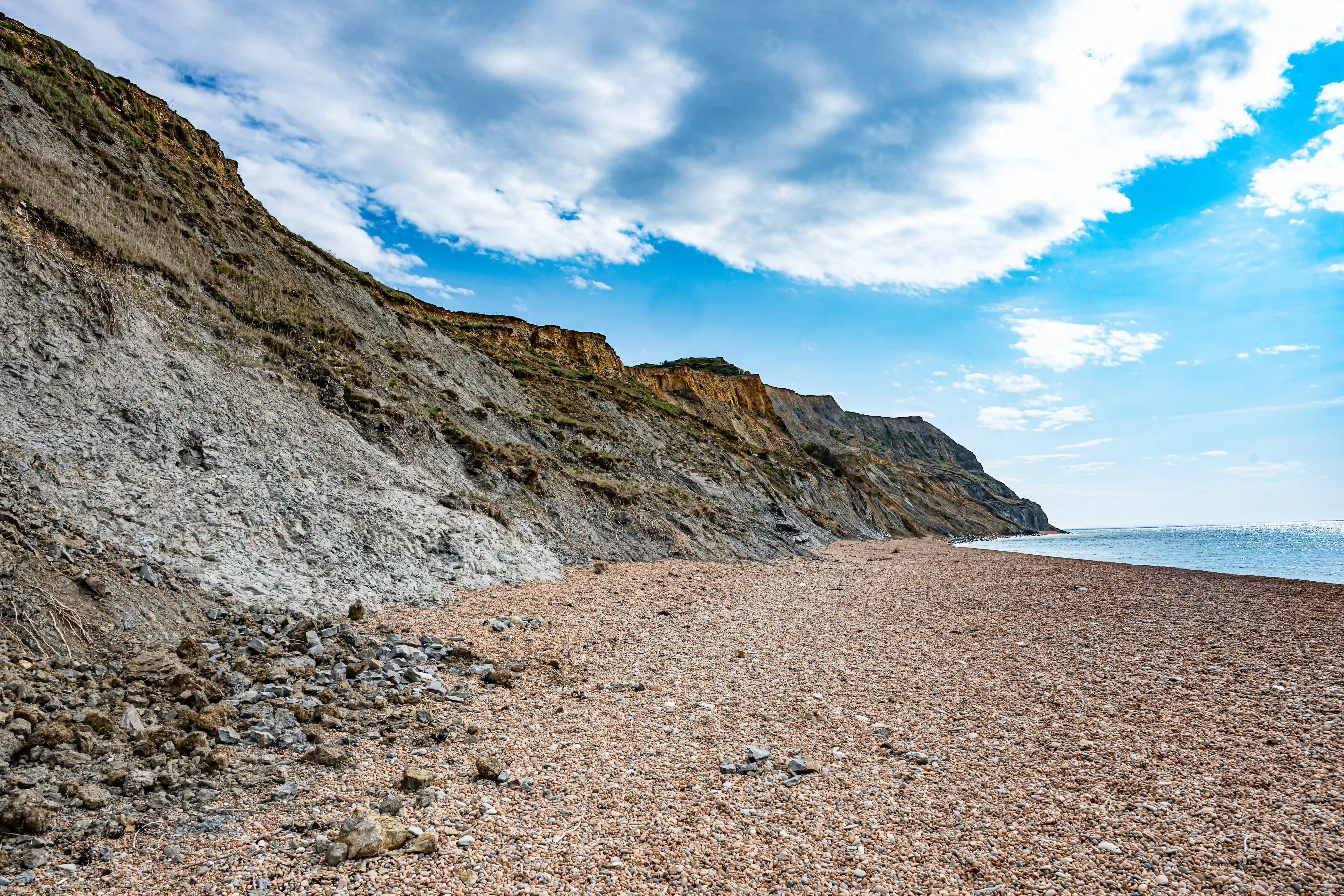 This image gives you a sense of walking along the coastline. The view is dominated by the imposing cliff face, which looks quite weathered and grey in parts, with layers of rock clearly visible. The pebbly beach stretches out ahead, and the sea continues on the right. The sky is a vibrant blue with dramatic white clouds, suggesting a pleasant, if perhaps breezy, day. It's a perspective that really highlights the scale of the natural landscape.