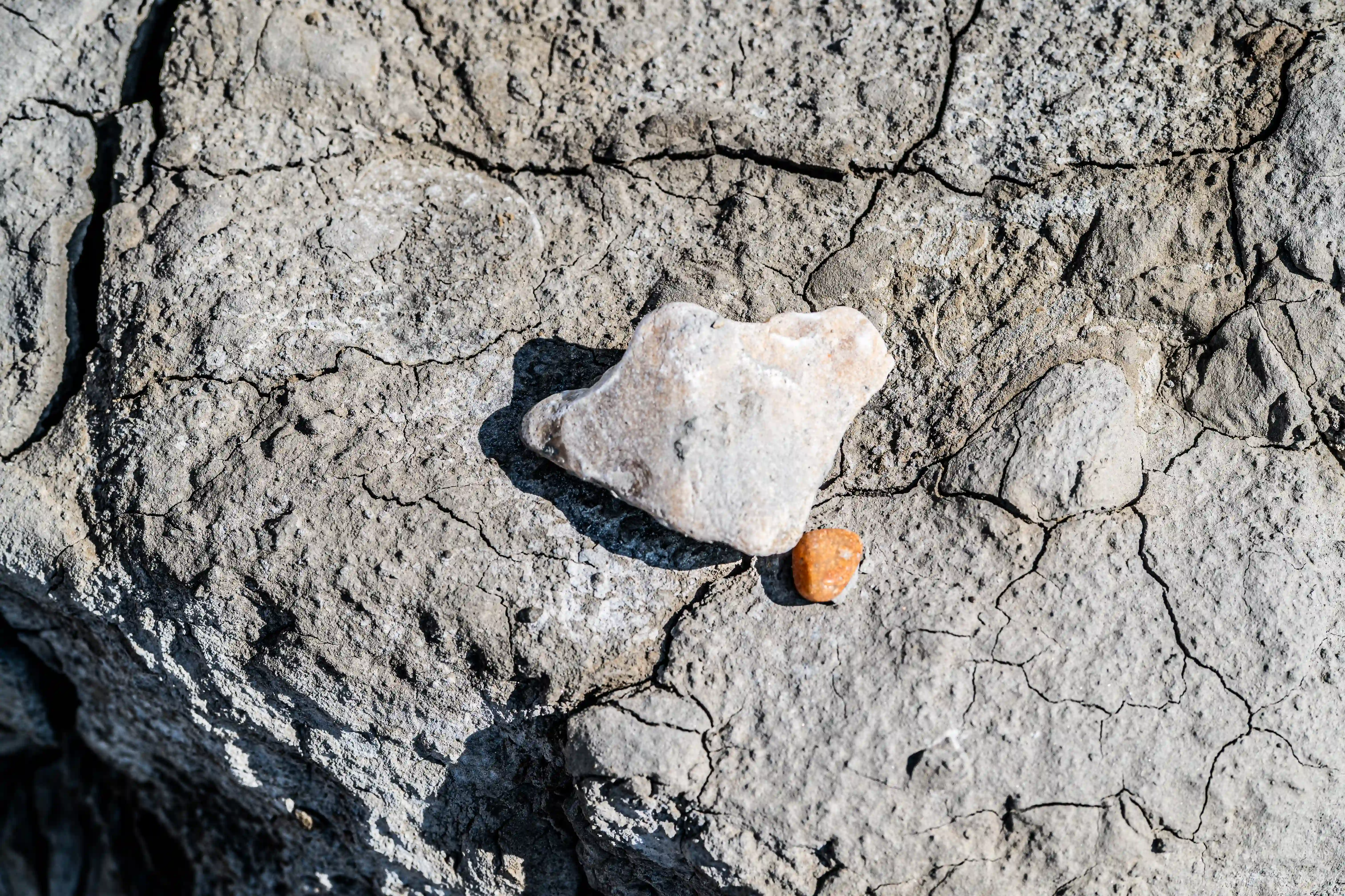 The image shows a cracked, dry surface, possibly indicating a desert or arid environment. On this surface, there is a heart-shaped rock and a small, round, orange object. The heart-shaped rock is light-colored and appears to be smooth, contrasting with the rough, cracked ground beneath it. The orange object is positioned near the tip of the heart shape, adding a pop of color to the otherwise muted tones of the scene.