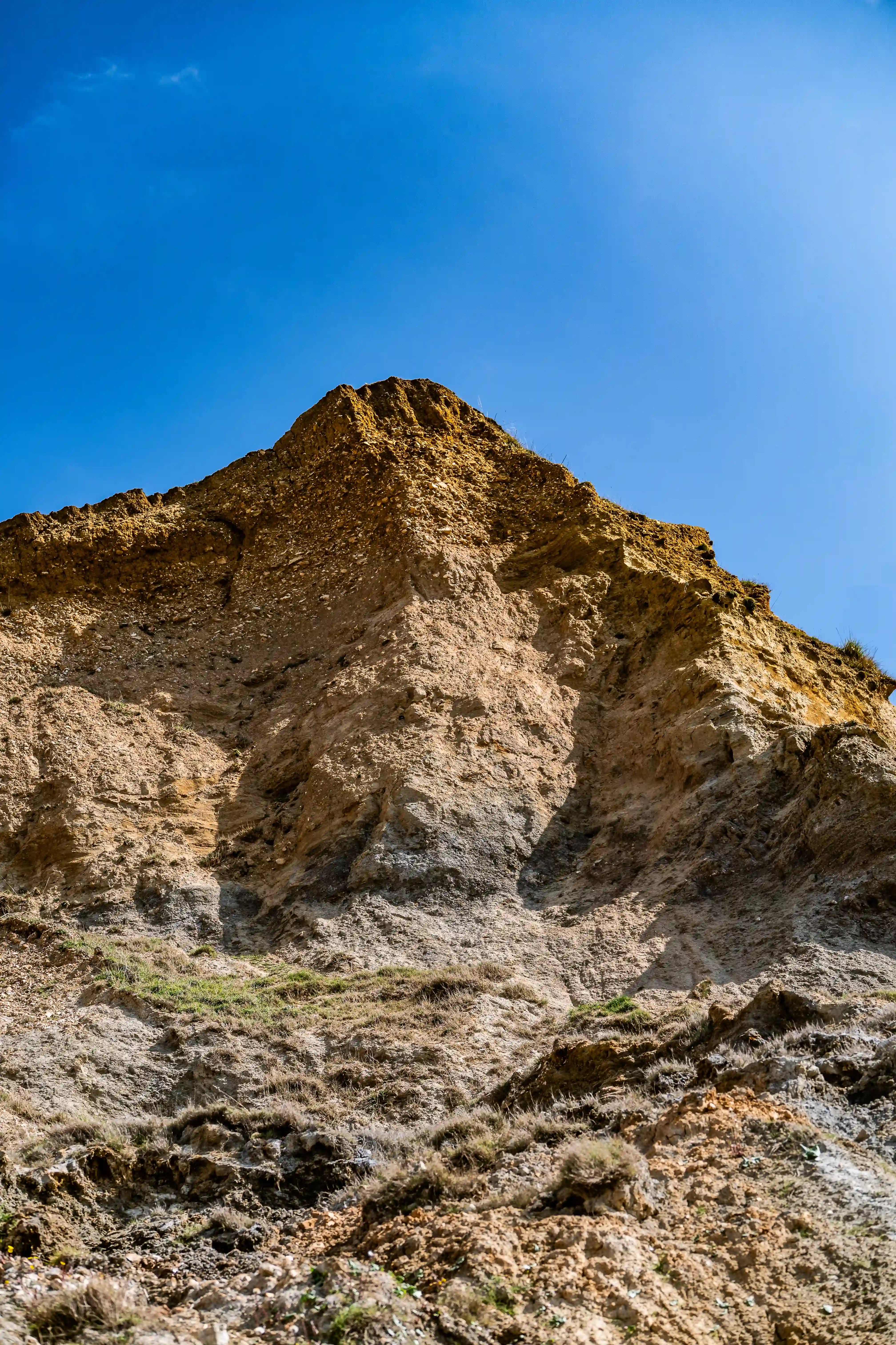 The image depicts a steep, rocky hillside with a clear blue sky in the background. The hillside is composed of light brown, layered rock formations with sparse vegetation at the base.