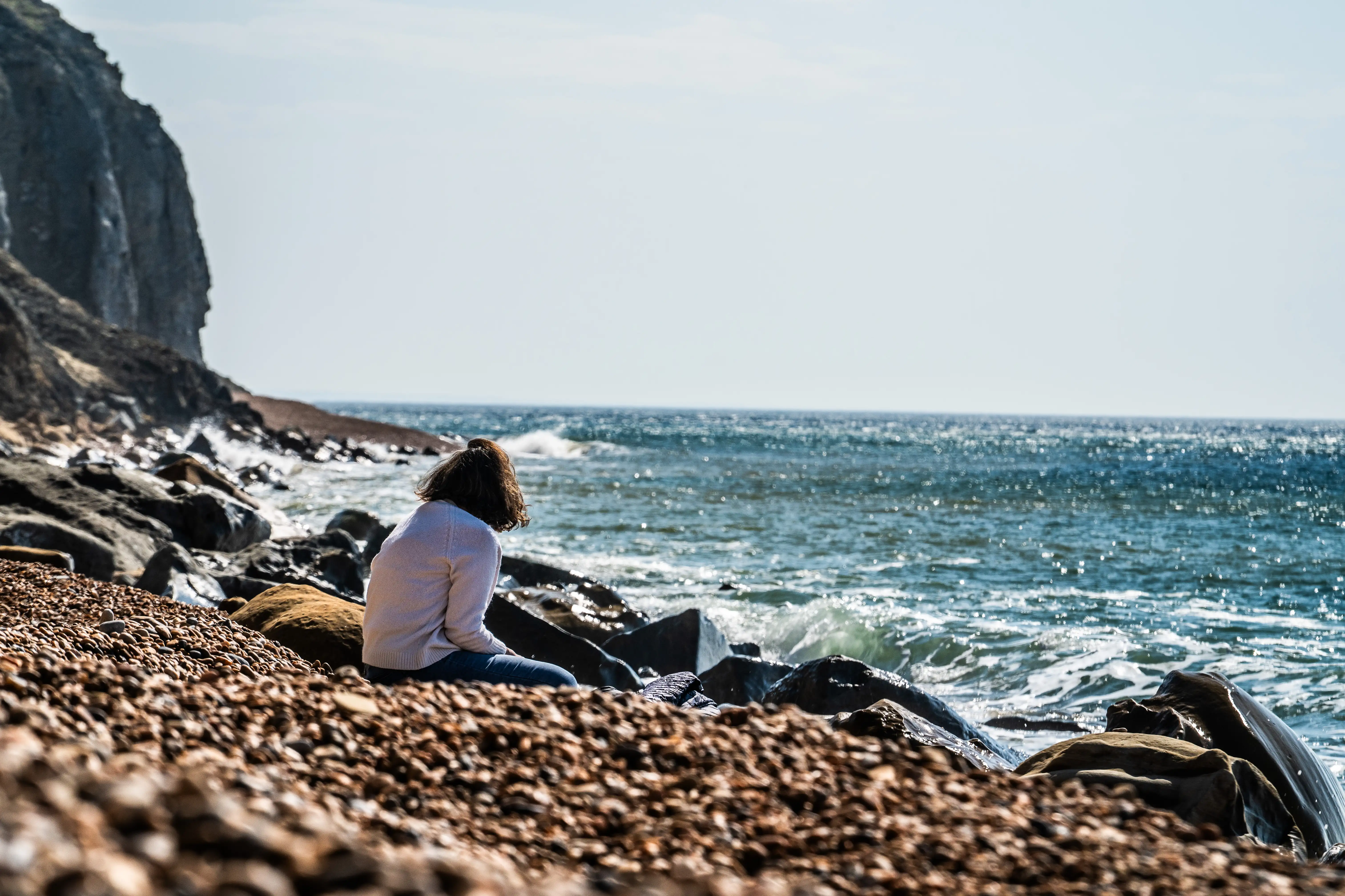 This picture shows a person, viewed from behind, sitting on a pebble beach and gazing out at the sea. The waves are crashing in the distance, and the water is a shimmering blue. The person is sitting amongst the stones and some larger rocks, looking quite peaceful and lost in thought. It's a serene scene, capturing a moment of quiet reflection by the coast.