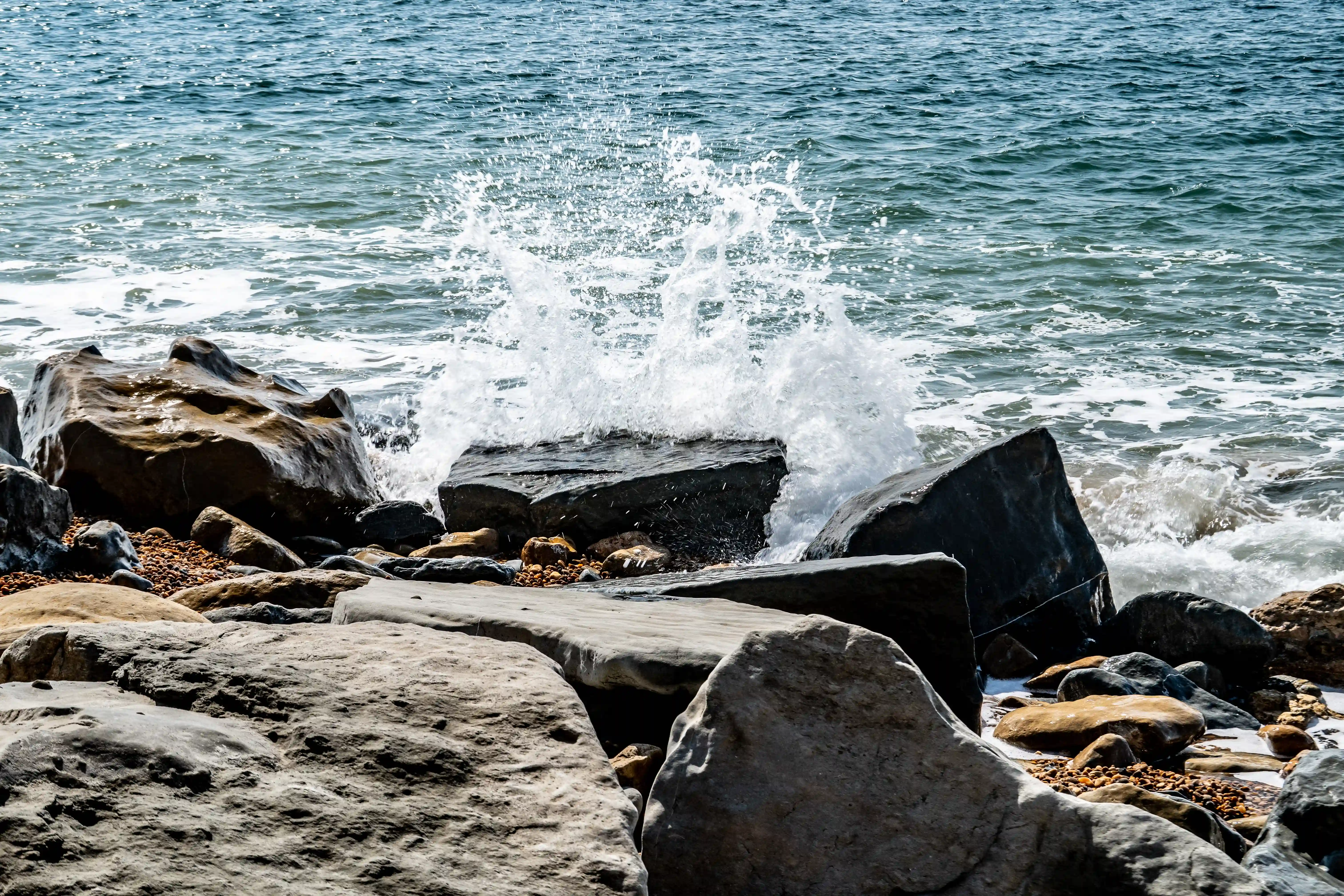 The image depicts a coastal scene with waves crashing against large rocks. The water is a deep blue-green, and the rocks are dark and jagged. The waves create splashes as they hit the rocks, capturing a dynamic and natural moment by the sea.
