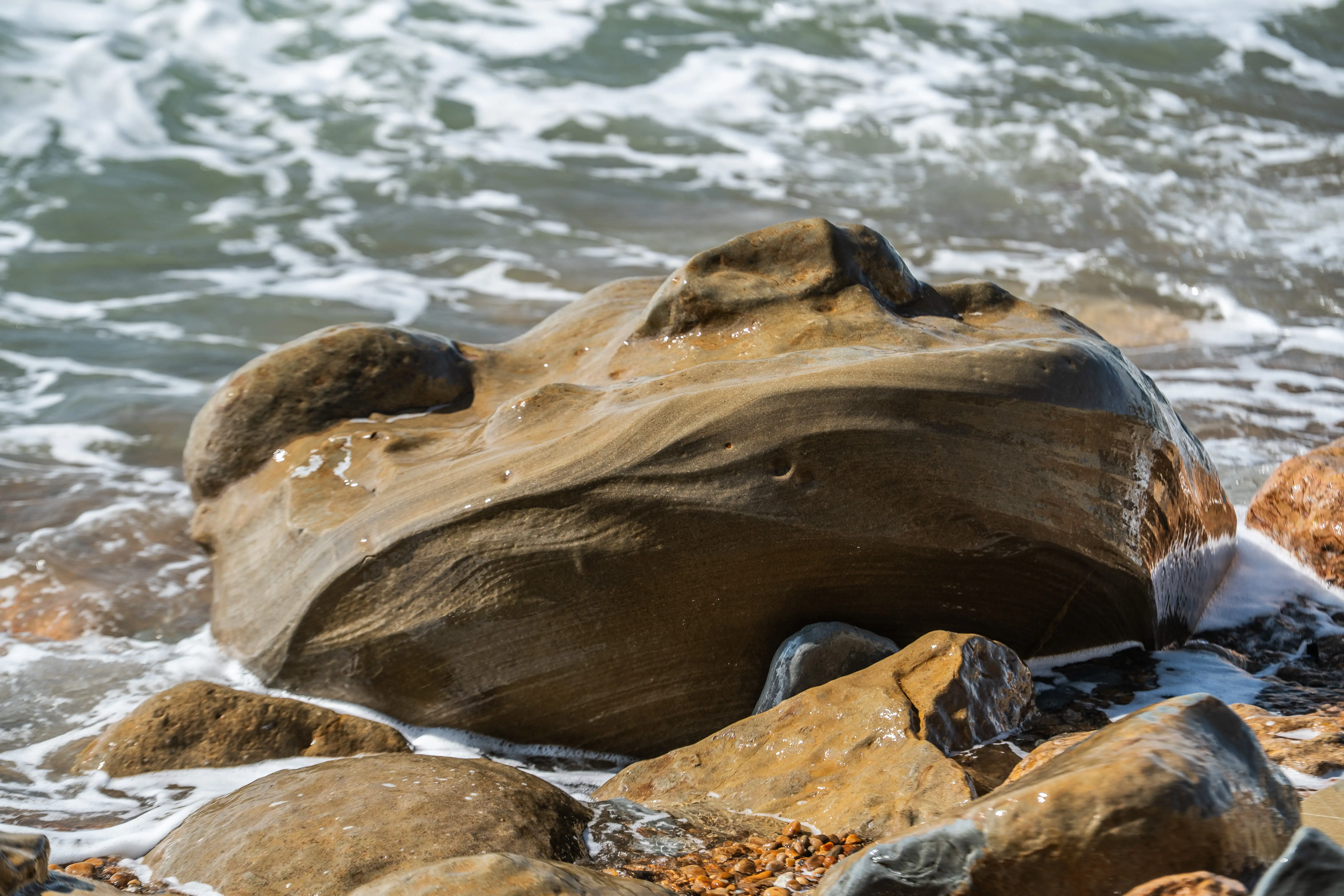 The image depicts a close-up view of a large, smooth rock formation situated near the edge of a body of water, possibly a sea or an ocean. The rock has a unique, almost helmet-like shape with a hollow center, and it is surrounded by smaller rocks and pebbles. The water is gently lapping against the rocks, creating a serene and natural scene.