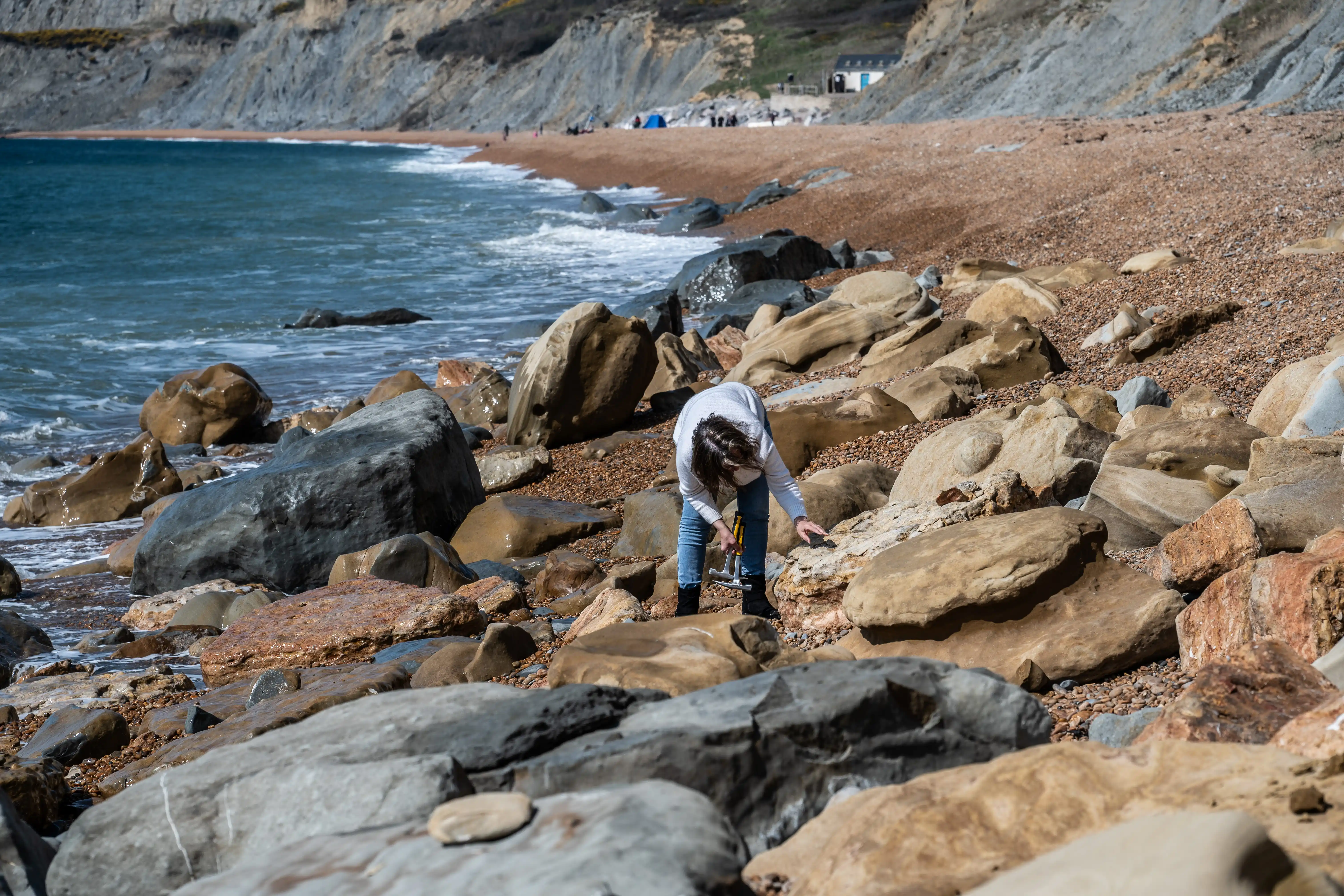The image depicts a person on a rocky beach, examining or collecting items among the rocks near the shoreline. The beach is surrounded by steep cliffs, and there are a few other people visible in the distance. The sea is calm, with gentle waves lapping against the rocks and pebbles.