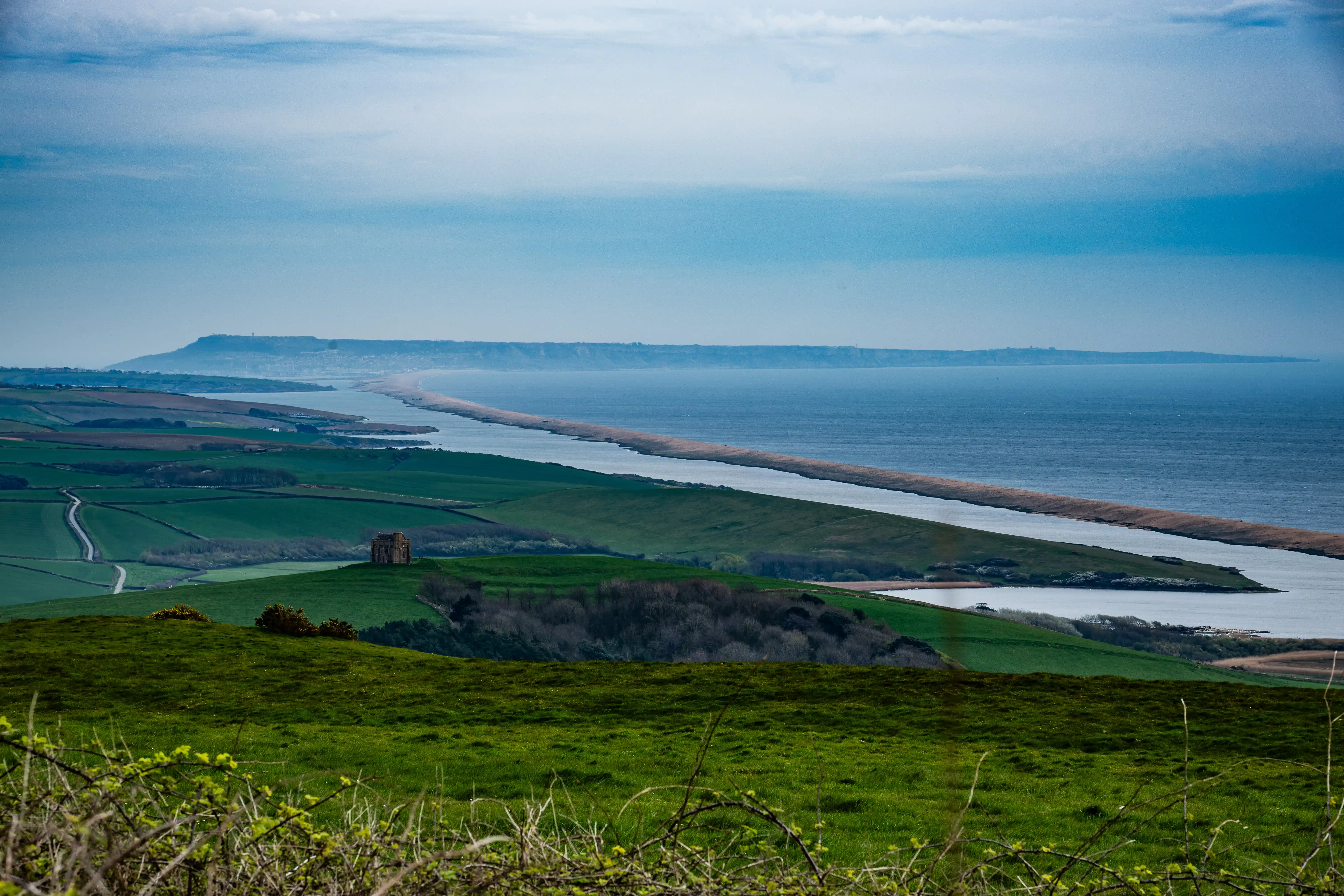 The image depicts a scenic landscape with a river flowing into the sea, surrounded by lush green fields and hills. The sky is overcast, and there is a prominent ruin or structure situated on one of the hills near the river.