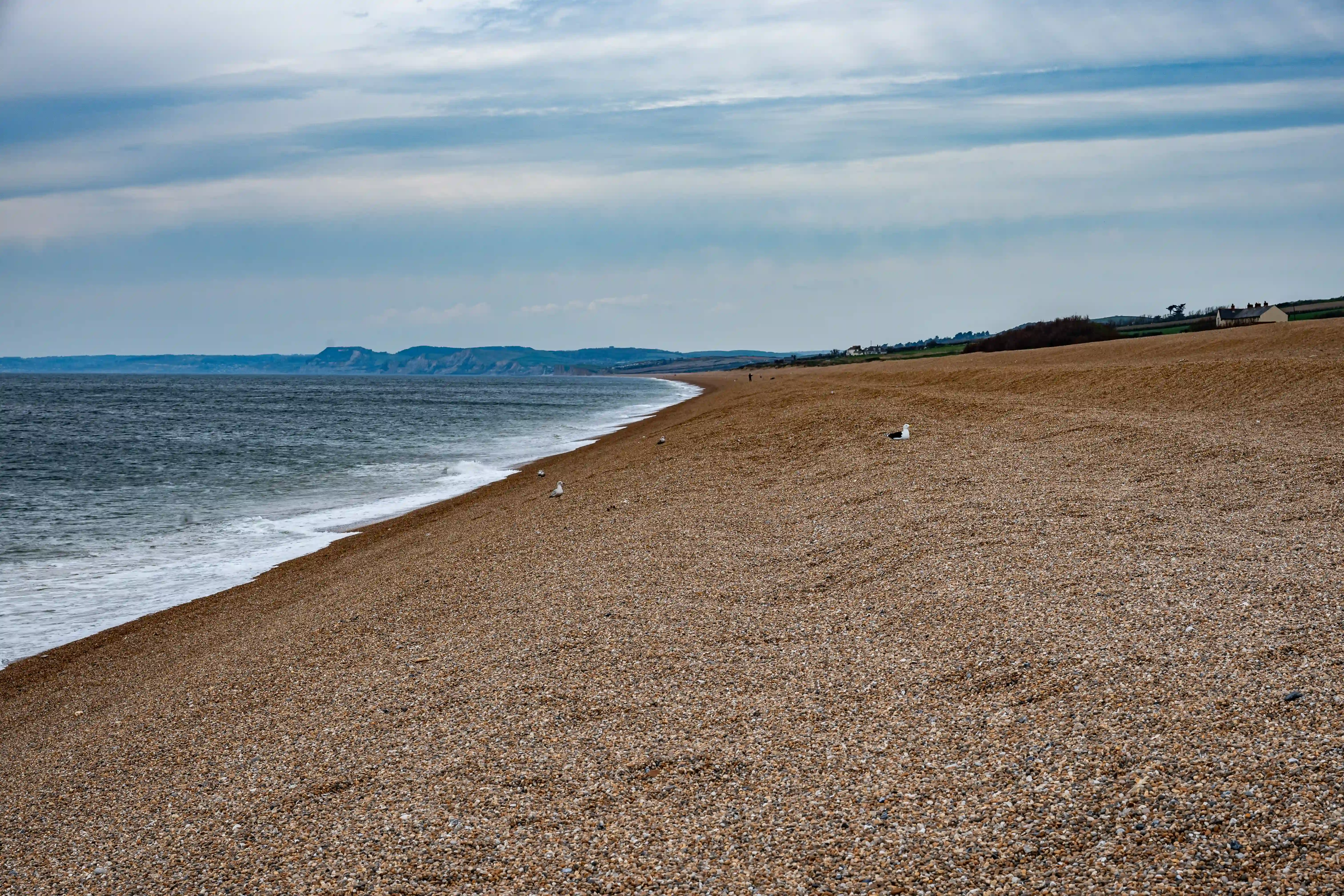 The image depicts a long, pebbly beach with the sea on the left side. The shoreline curves gently, leading to distant cliffs and a slightly cloudy sky overhead. The beach appears mostly deserted, with a few scattered objects and a lone figure in the distance.