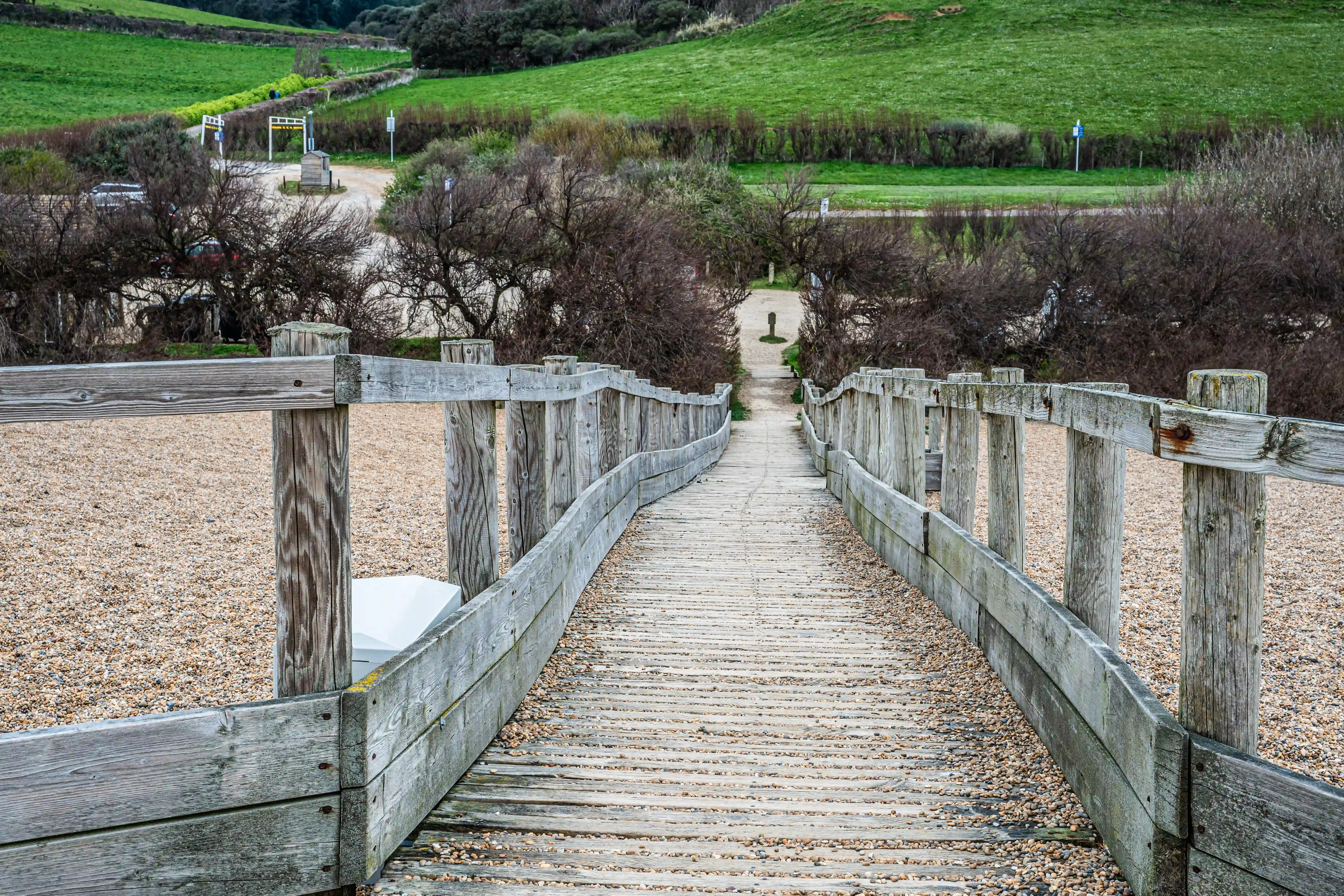 The image depicts a wooden walkway with railings on both sides, leading through a natural landscape. The path is surrounded by a mix of green grassy hills and leafless trees, suggesting it might be late autumn or early spring. The walkway appears to be well-maintained and leads towards a distant point that is not clearly visible. The surrounding area includes a dirt road with a few vehicles and some signage in the background, indicating it might be near a parking area or an entrance to a natural reserve or park.