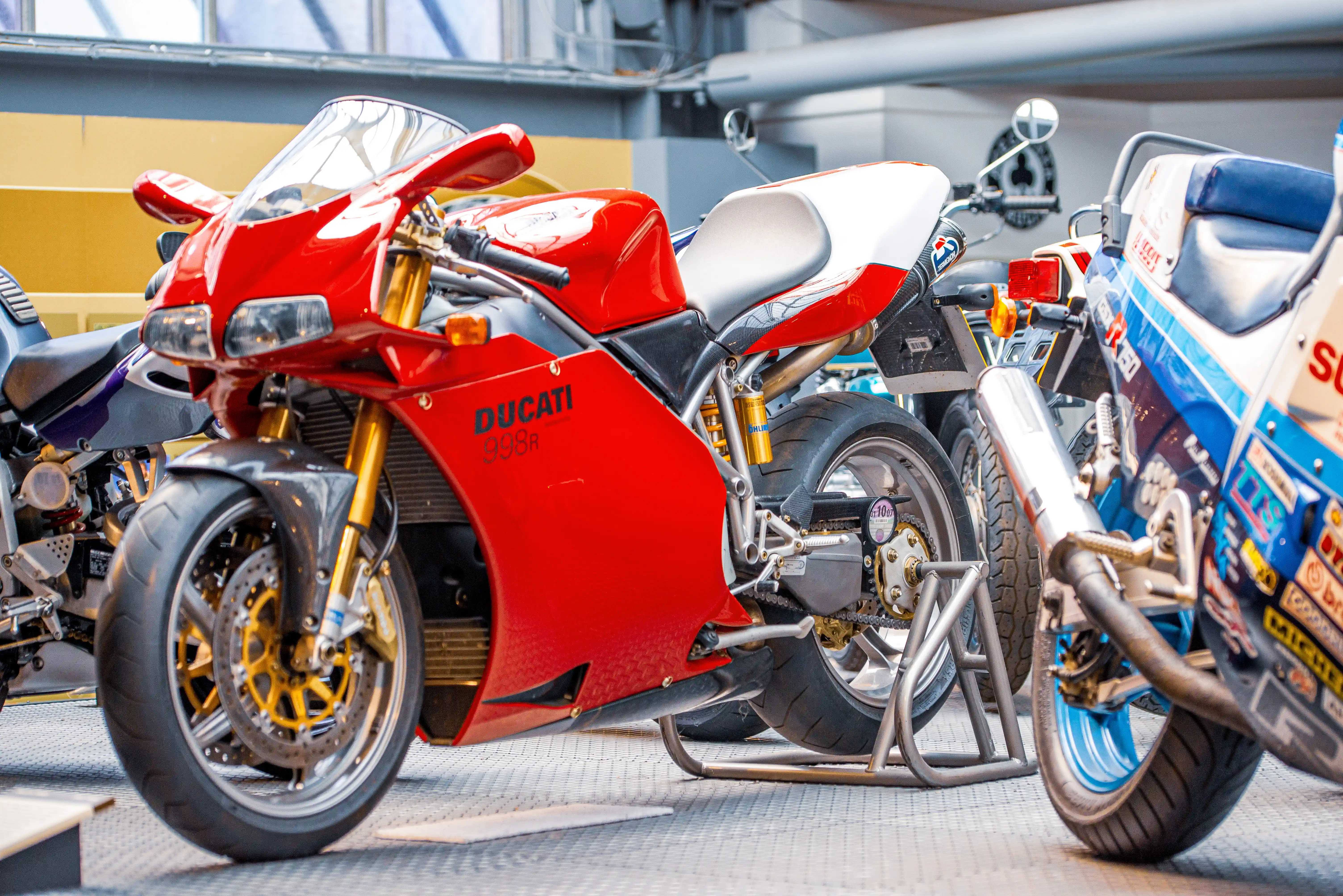 The image shows three motorcycles parked side by side. The motorcycle in the foreground is a red Ducati 958r with a distinctive red fairing and gold-colored components. The other two motorcycles have different designs and colors, with one featuring a predominantly white and blue color scheme and the other being black. The motorcycles are parked in what appears to be a garage or storage area with a concrete floor and metal beams in the background.