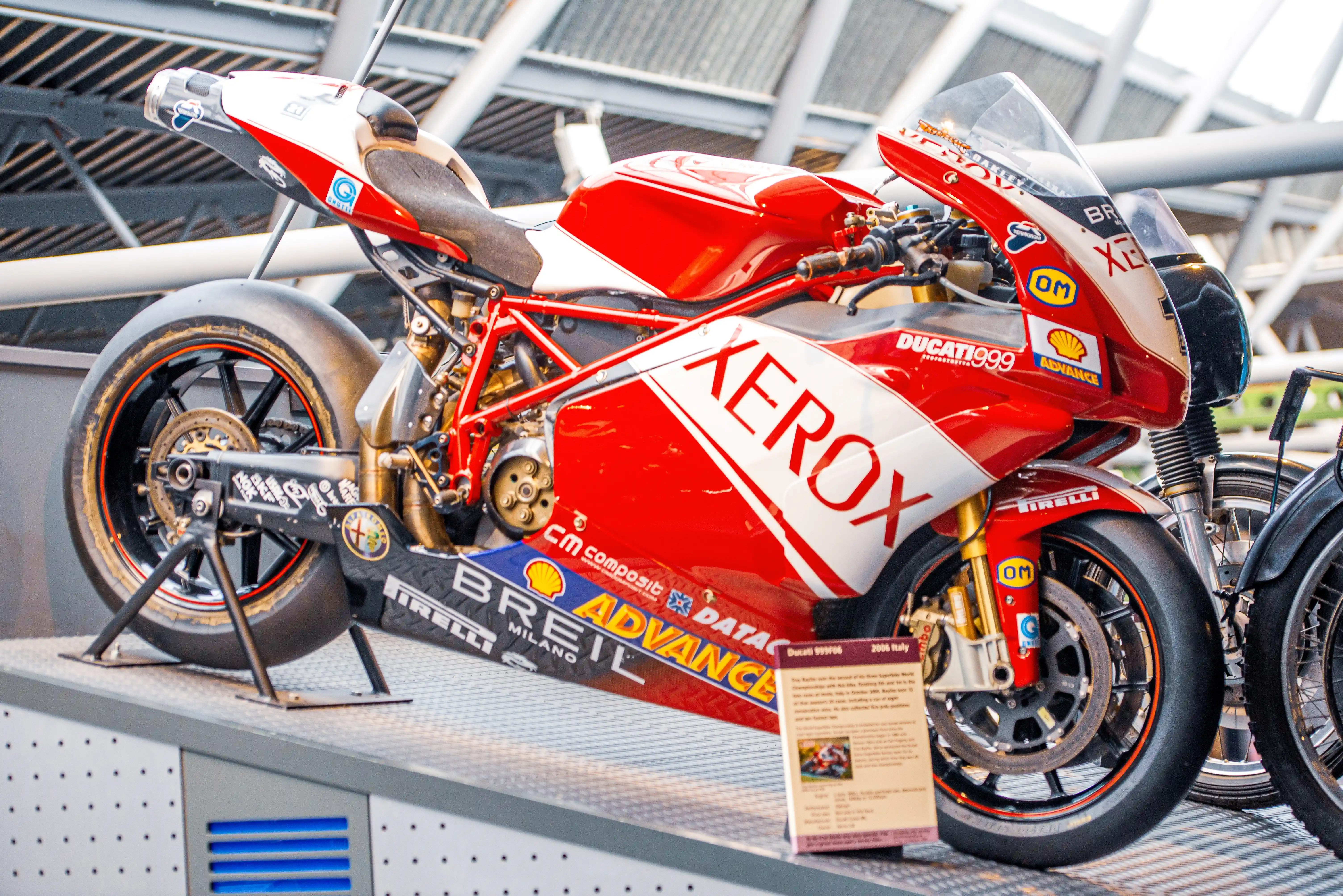 The image showcases a Ducati 999 motorcycle, prominently featuring the Zerox brand. The motorcycle is part of a display, likely in a museum or exhibition setting, as indicated by the informational plaque next to it. The bike is equipped with various sponsor logos, including Shell, Pirelli, and others, and has a distinctive red and white color scheme. The motorcycle appears to be a racing model, highlighted by its sleek design and exposed mechanical components.