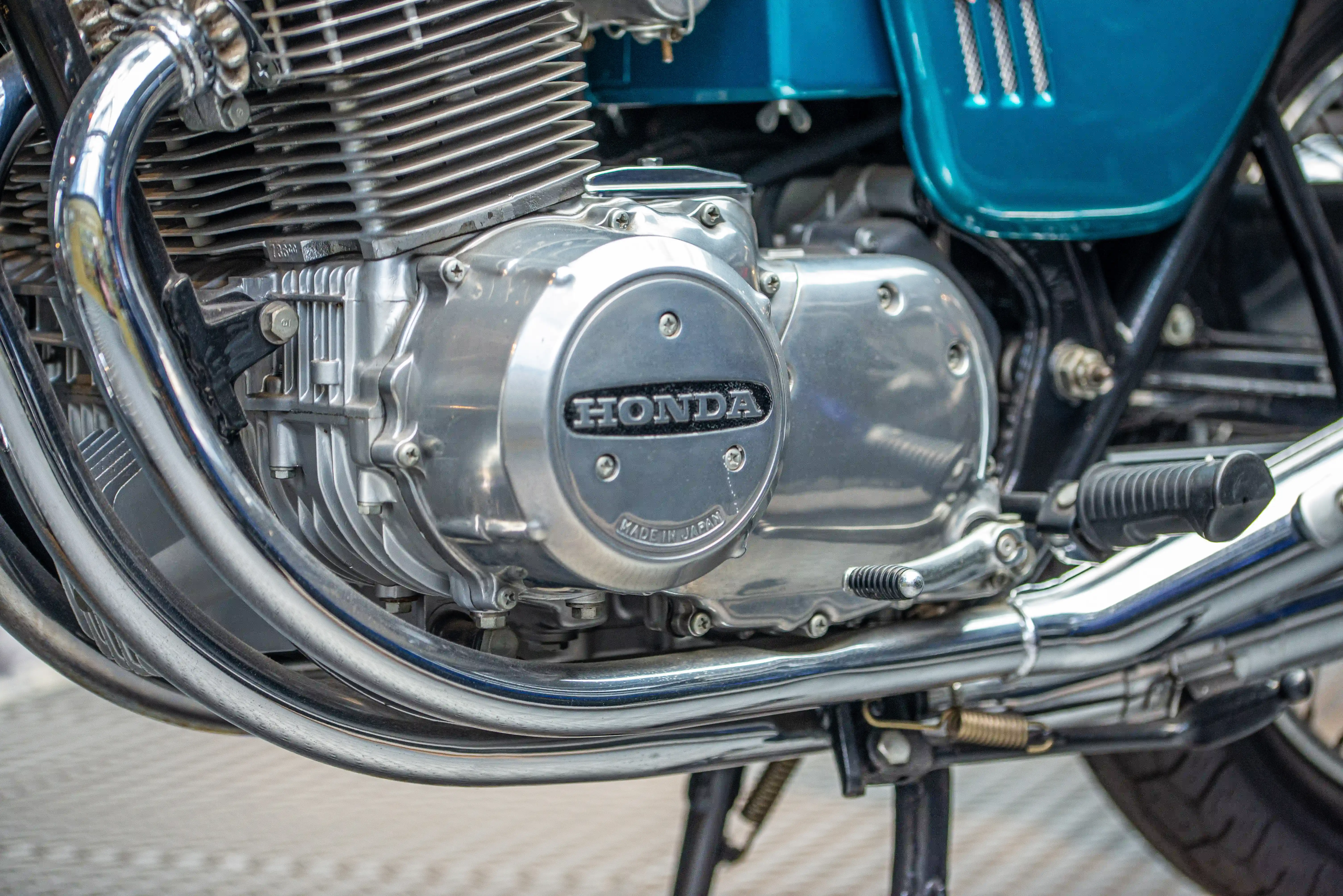 The image shows a close-up view of a motorcycle engine, prominently featuring the Honda brand logo on the engine cover. The motorcycle appears to be well-maintained and has a classic design, with visible components such as the cylinder head, fins, and various bolts and nuts. The engine cover also indicates that it is made in Japan. The surrounding area includes parts of the motorcycle's frame and some wiring.