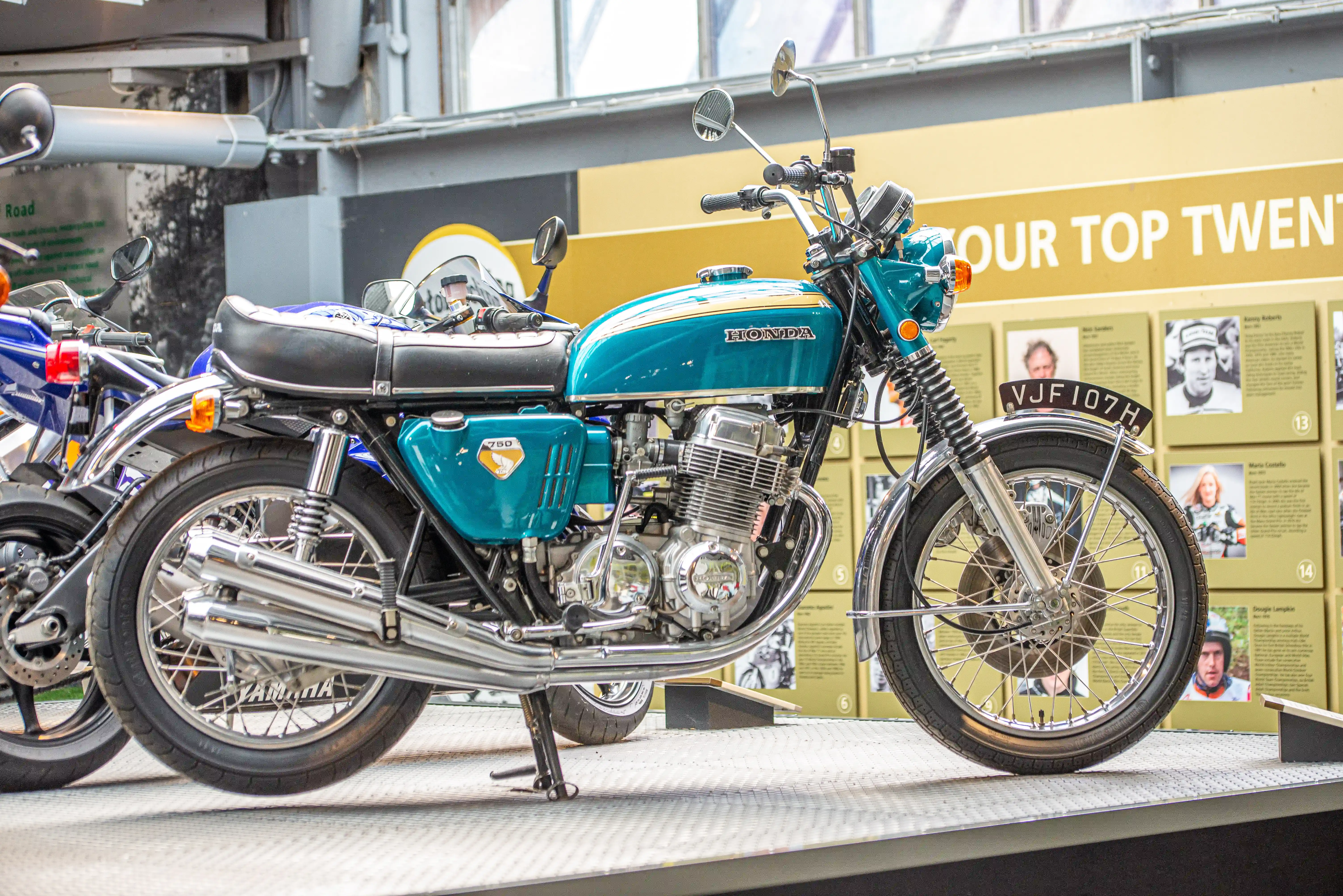 The image showcases a classic Honda motorcycle, specifically a model 750, displayed in a museum or exhibition setting. The motorcycle is teal and black with chrome details, and it features a distinctive design typical of older models. It is positioned on a platform with other motorcycles and informational displays in the background, highlighting notable individuals and their contributions. The setting suggests a celebration of motorcycle history and innovation.