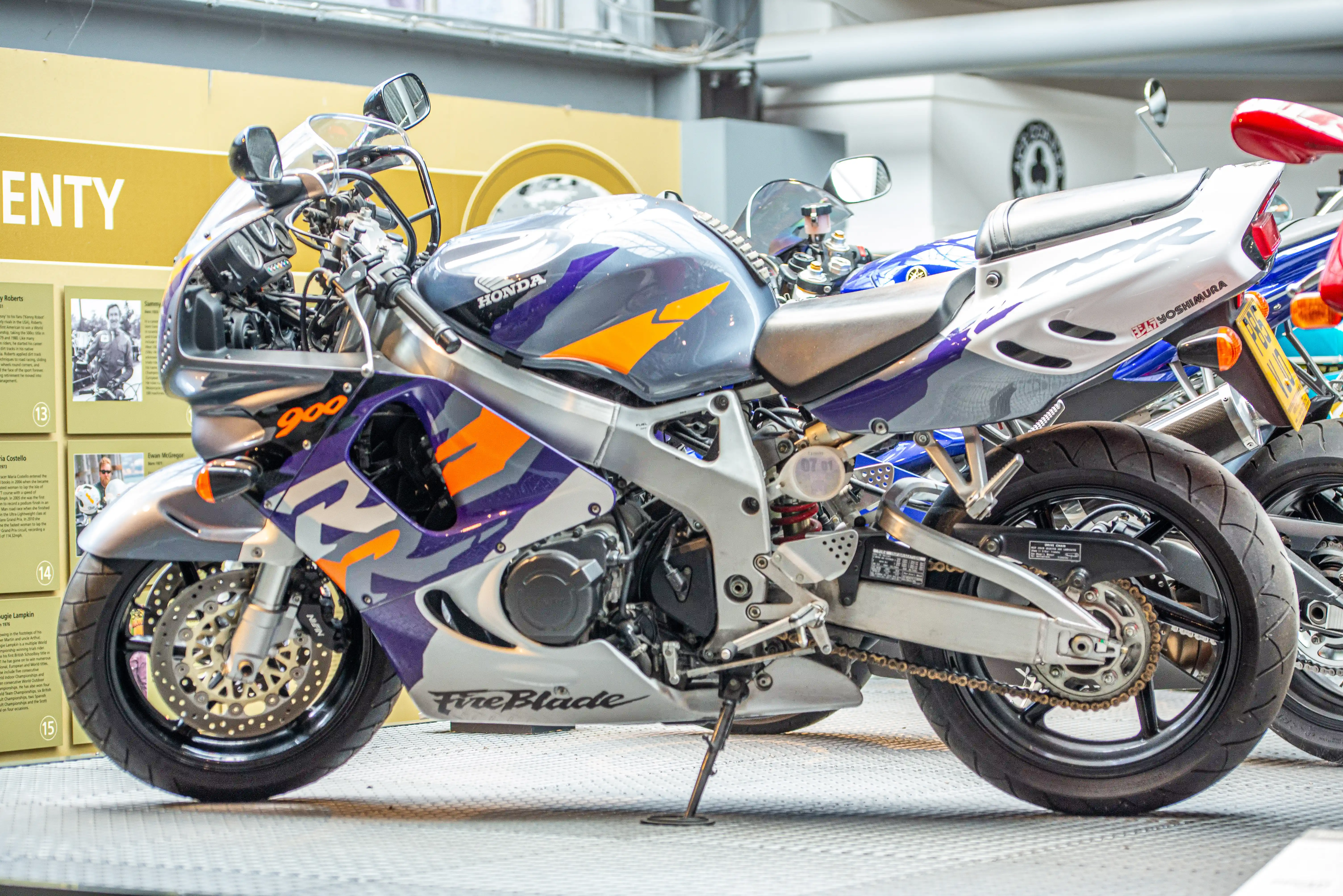 Right then, this first one is a proper beauty! It's a Honda CBR900RR Fireblade, and it looks like it's in absolutely mint condition. You can see the iconic 'Fireblade' logo proudly displayed on the fairing. It's got a wicked silver and purple paint job with some nice orange accents that really make it pop. The bike's a bit of a legend, really, known for its performance and how light it felt for its size back in the day. It's sitting on a display stand, looking ready to tear up the road, or maybe it's in a museum somewhere, judging by the display boards in the background. Definitely a sporty number.
