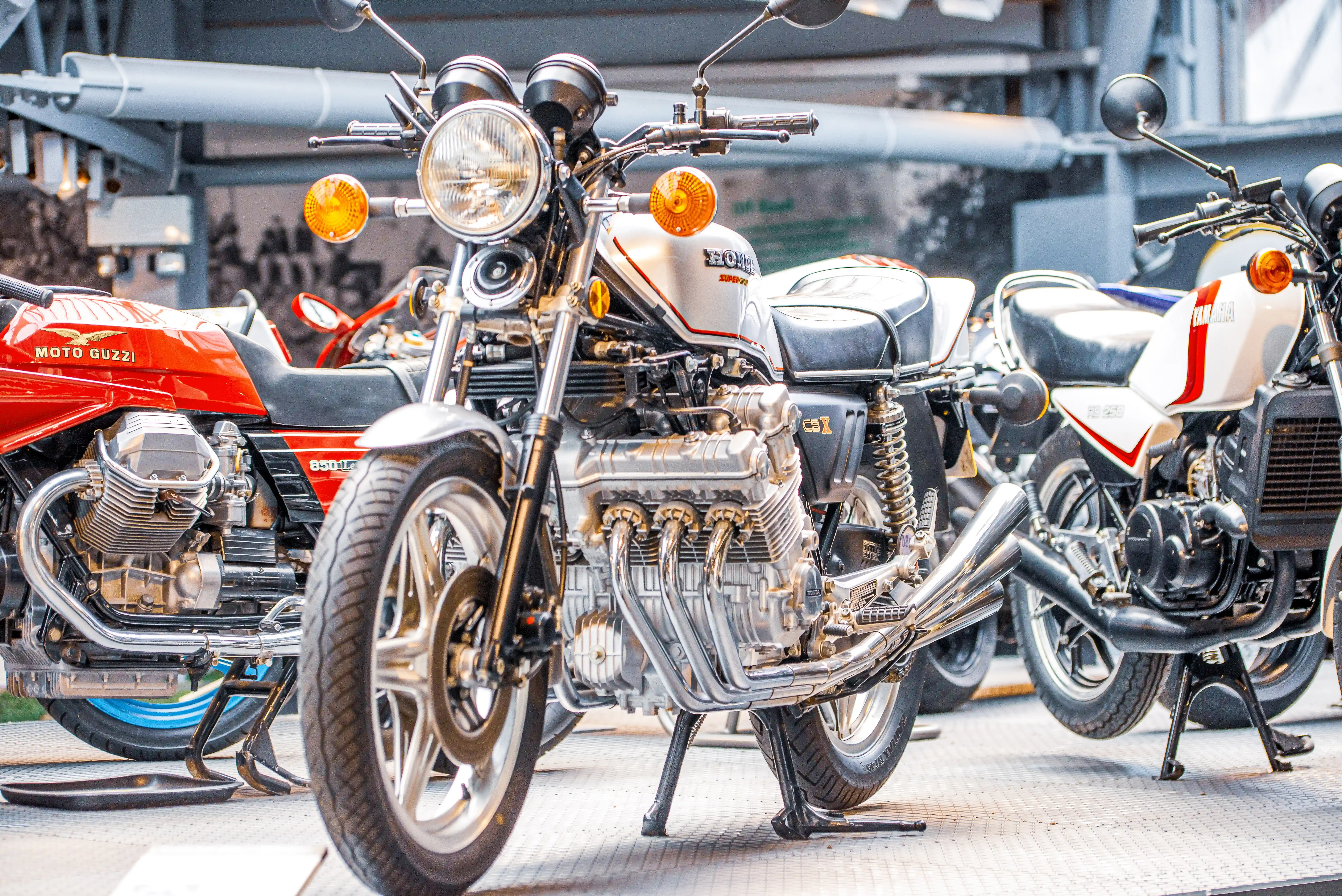 Now, this second image is a bit of a different vibe. We've got a few classic bikes lined up here, and the star of the show, right in the middle, is a Honda CBX. Look at that engine – it's a beast with six cylinders all lined up like soldiers! It's got that retro chrome look that's just timeless. To the left, you can spot a red Moto Guzzi, and to the right, another Honda, possibly a Gold Wing or something similar. These bikes have that old-school charm, all about that smooth ride and a bit of rumble. They look like they're ready for a leisurely cruise, perhaps down the coast or to a classic bike rally. It’s a real collection of motorcycling history here.