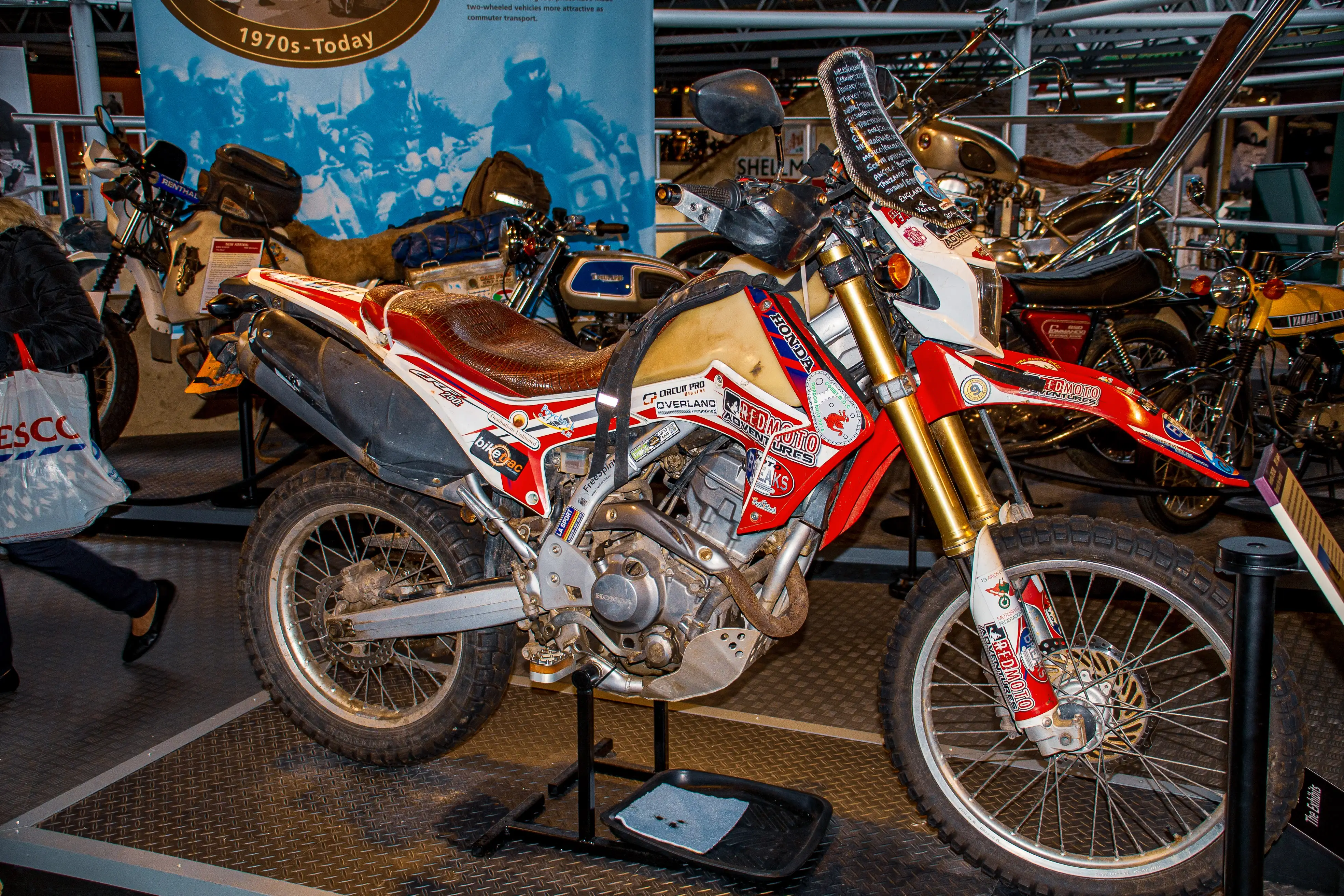 he image showcases a collection of motorcycles on display, with a prominent red, white, and gold motorcycle in the foreground. This motorcycle features various stickers and sponsors' logos, including KTM, Red Bull, and Mitas, and is equipped with a leather seat and gold-colored front forks. The background includes other motorcycles and a banner indicating the exhibit covers motorcycles from the 1970s to the present day. The setting appears to be an indoor exhibition space, likely a museum or showroom, with various motorcycle parts and accessories visible.