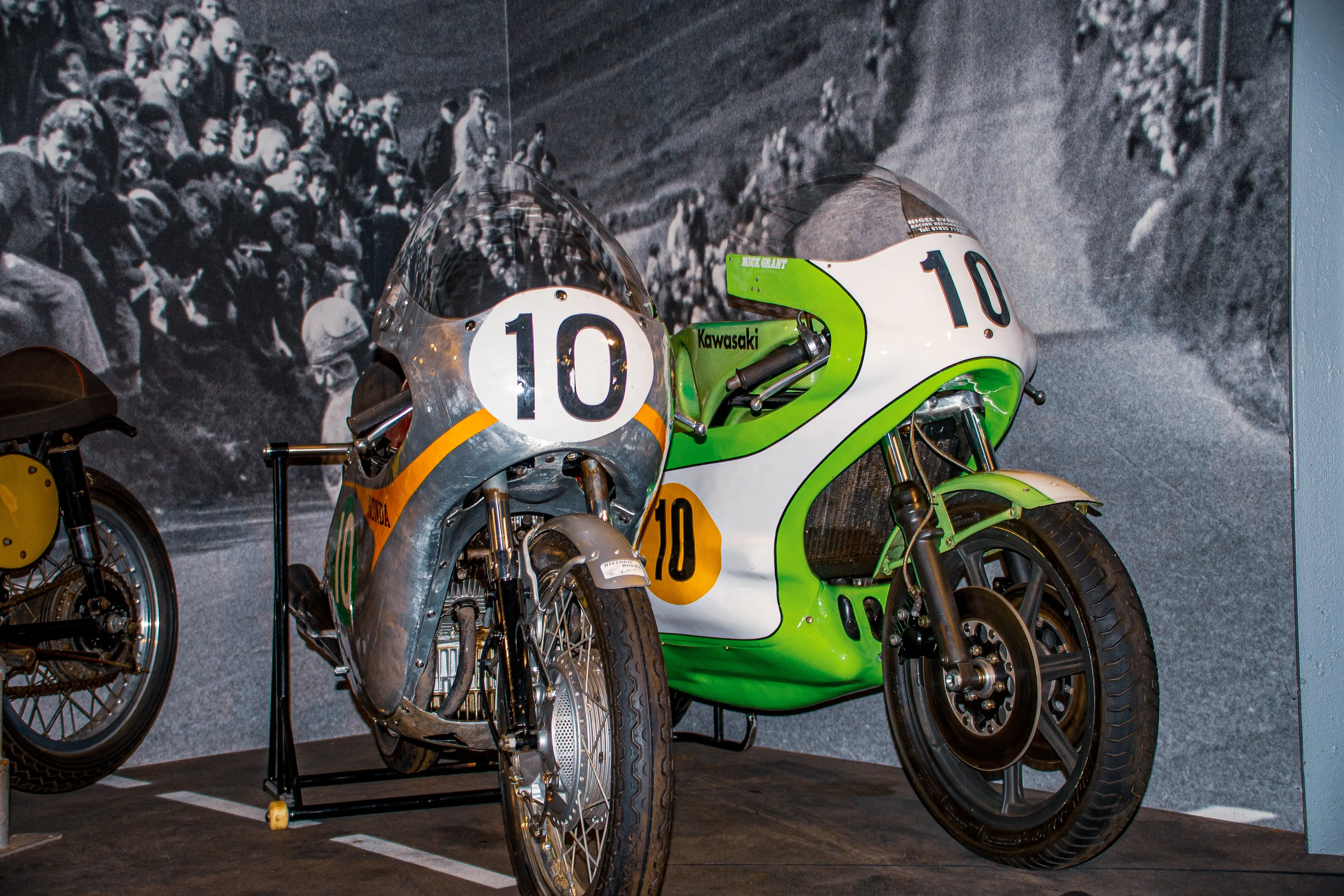 The image shows two vintage racing motorcycles on display. The motorcycle in the foreground is a Kawasaki, painted in green, white, and orange, with the number 10 prominently displayed. The motorcycle in the background is yellow and black, with the number 10 as well. Both motorcycles are positioned on stands, and the background features a black-and-white photograph of a crowd, likely from a past racing event.