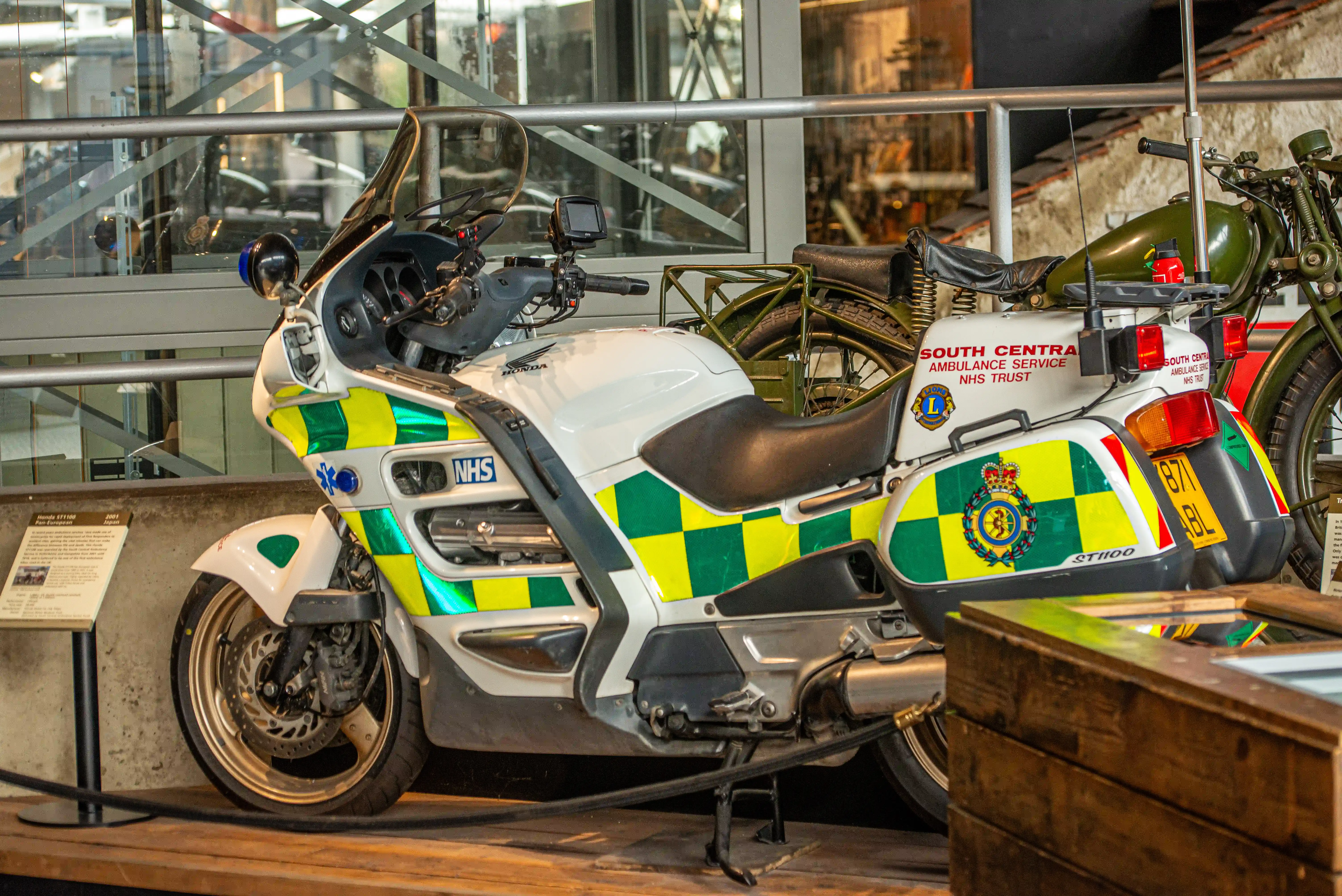 The image depicts an ambulance service motorcycle from the South Central Ambulance Service NHS Trust. The motorcycle is a Honda ST1100, equipped with medical supplies and marked with the NHS logo and checkered patterns in green and yellow. It is displayed in a museum-like setting, alongside another vintage motorcycle.