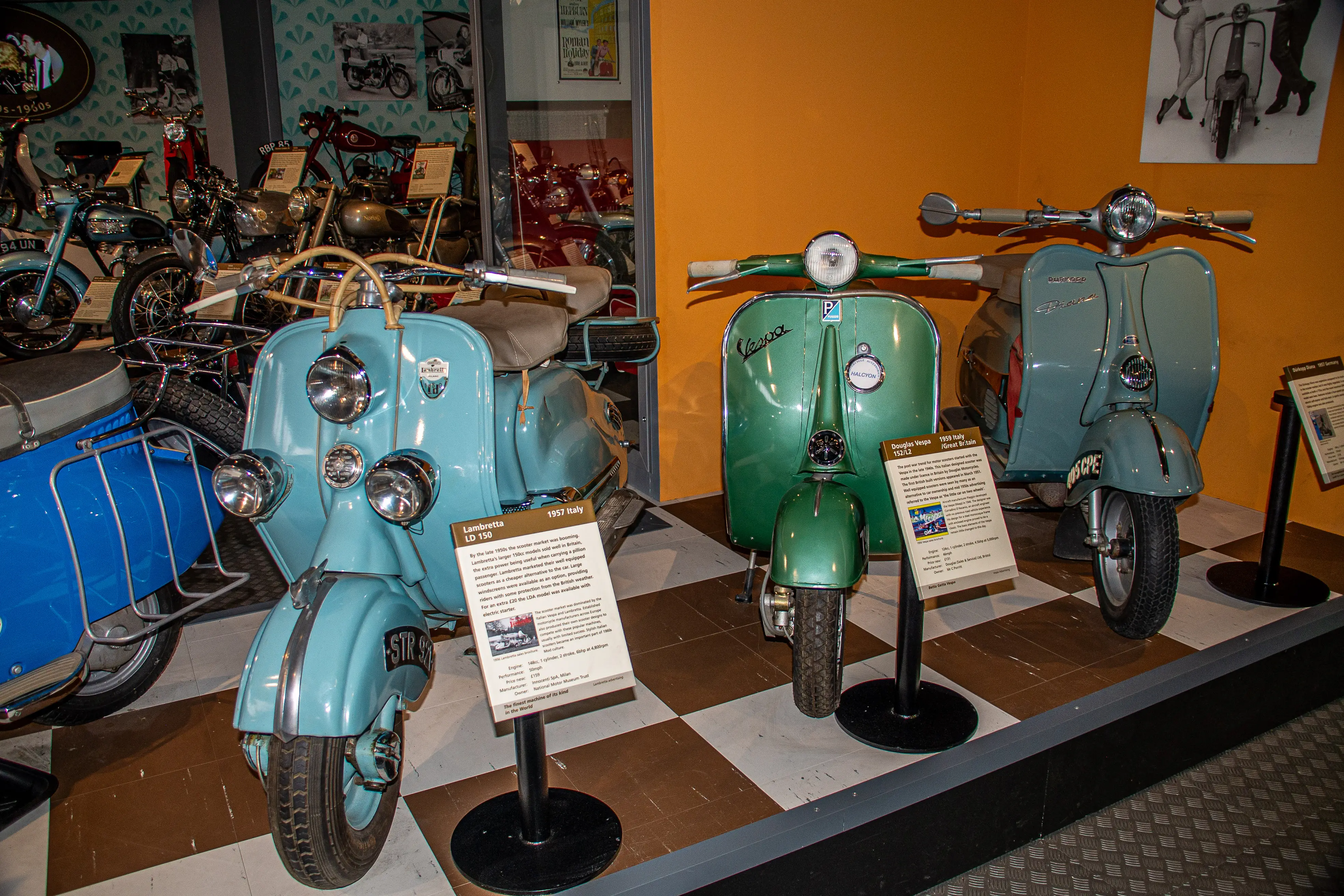 The image shows a display of vintage scooters in a museum setting. There are three scooters prominently featured: a Lambretta LD 150 from 1957 Italy and two Vespa models, including a Douglas Vespa 152 12 from 1953. Each scooter has an informational placard in front of it, providing details about its history and specifications. The background includes additional motorbikes and various vintage advertisements and posters related to scooters.
