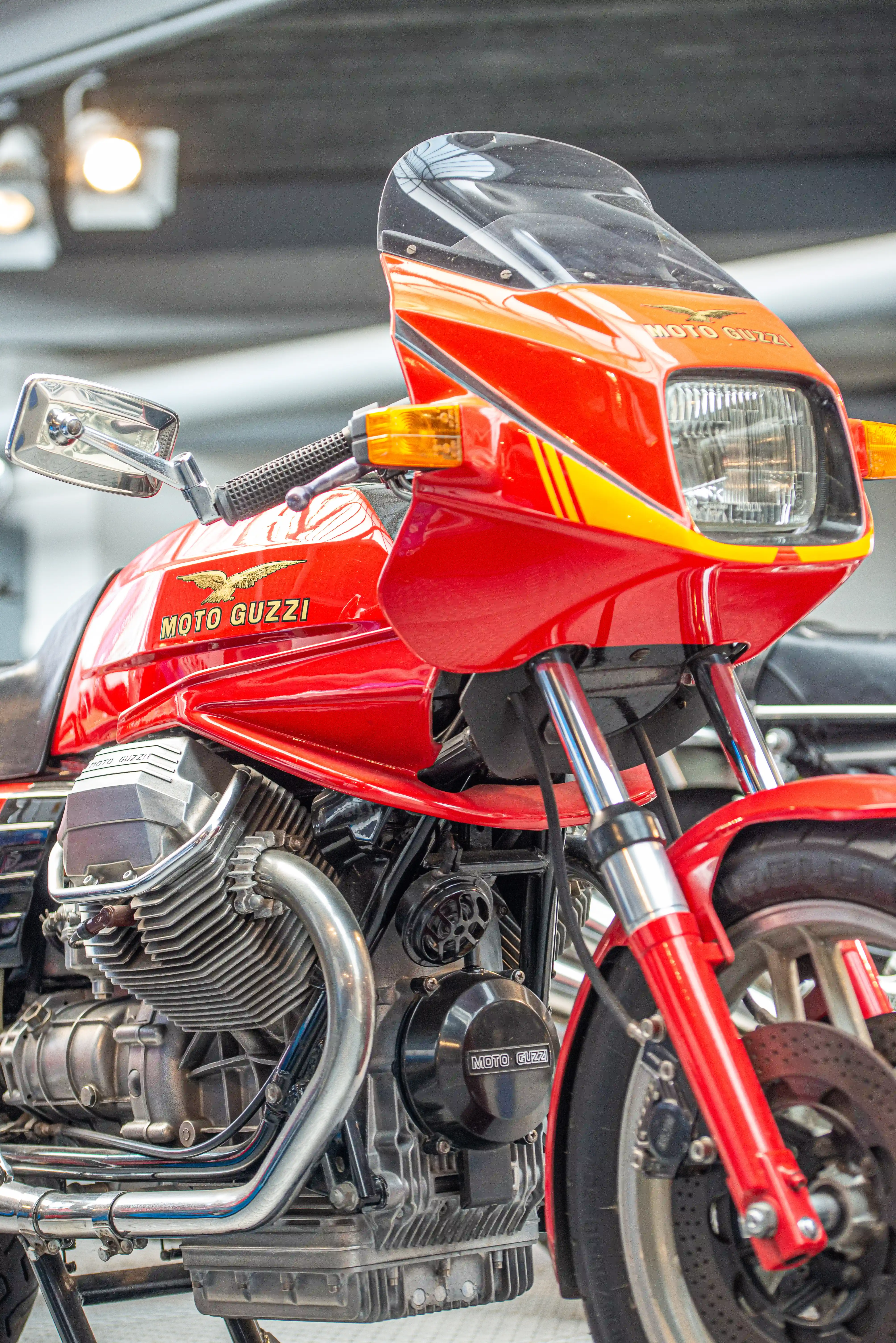 The image showcases a red Moto Guzzi motorcycle with a distinctive design, featuring a prominent front fairing, dual headlights, and a visible engine with the brand name 'Moto Guzzi' emblazoned on it. The motorcycle is parked indoors, possibly in a garage or showroom, with a sleek and polished appearance.