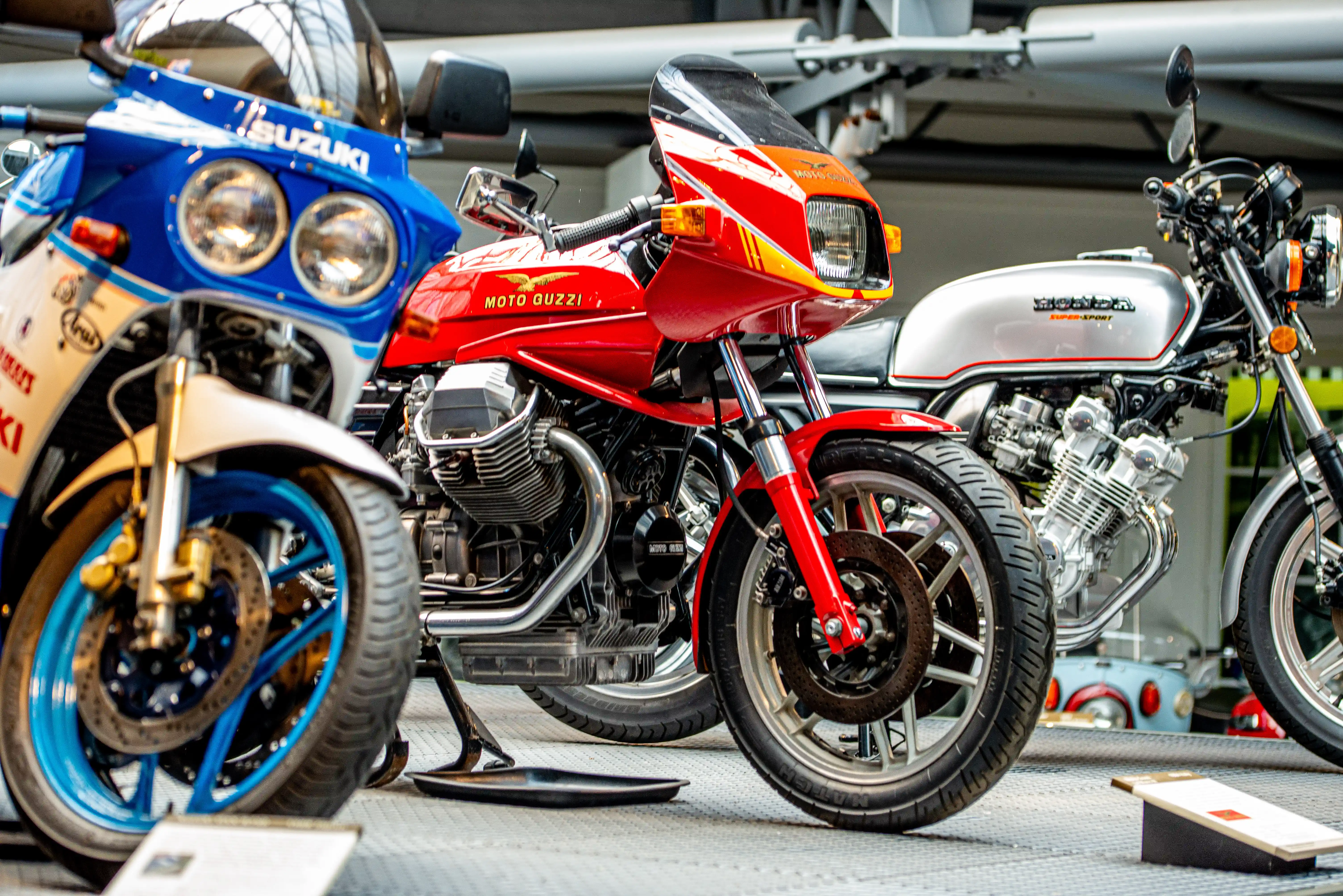 The image showcases three classic motorcycles parked side by side. The motorcycles are from different brands: Suzuki, Moto Guzzi, and Honda. Each motorcycle has distinct colors and designs, with the Suzuki being blue, the Moto Guzzi being red, and the Honda being silver. They are displayed in what appears to be a garage or showroom setting, with various tools and equipment visible in the background