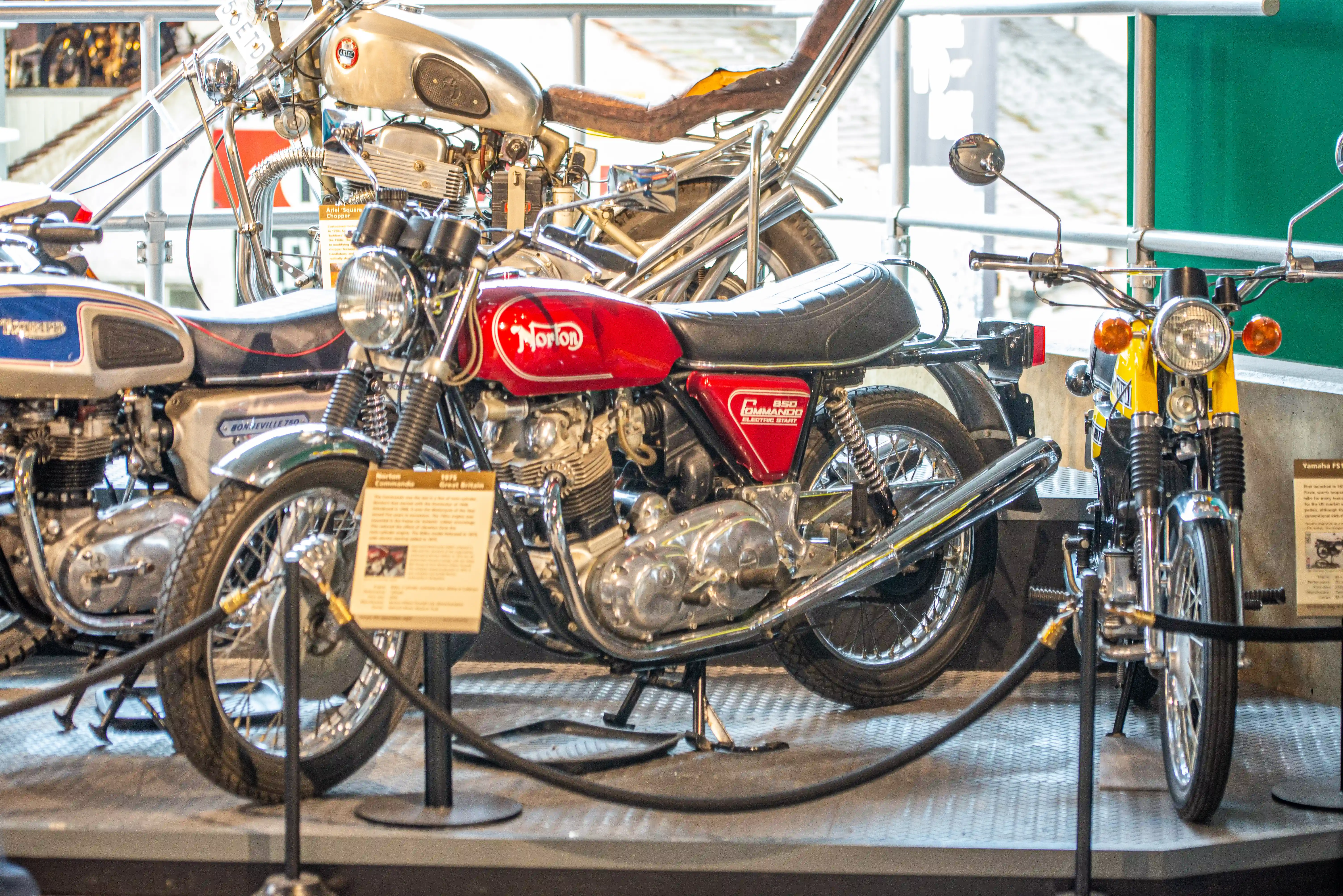 The image shows a collection of vintage motorcycles on display, including a prominent Norton motorcycle in the center with a red fuel tank and chrome engine, flanked by other classic bikes. Each motorcycle is mounted on stands with informational placards in front of them, suggesting they are part of an exhibition or museum display.