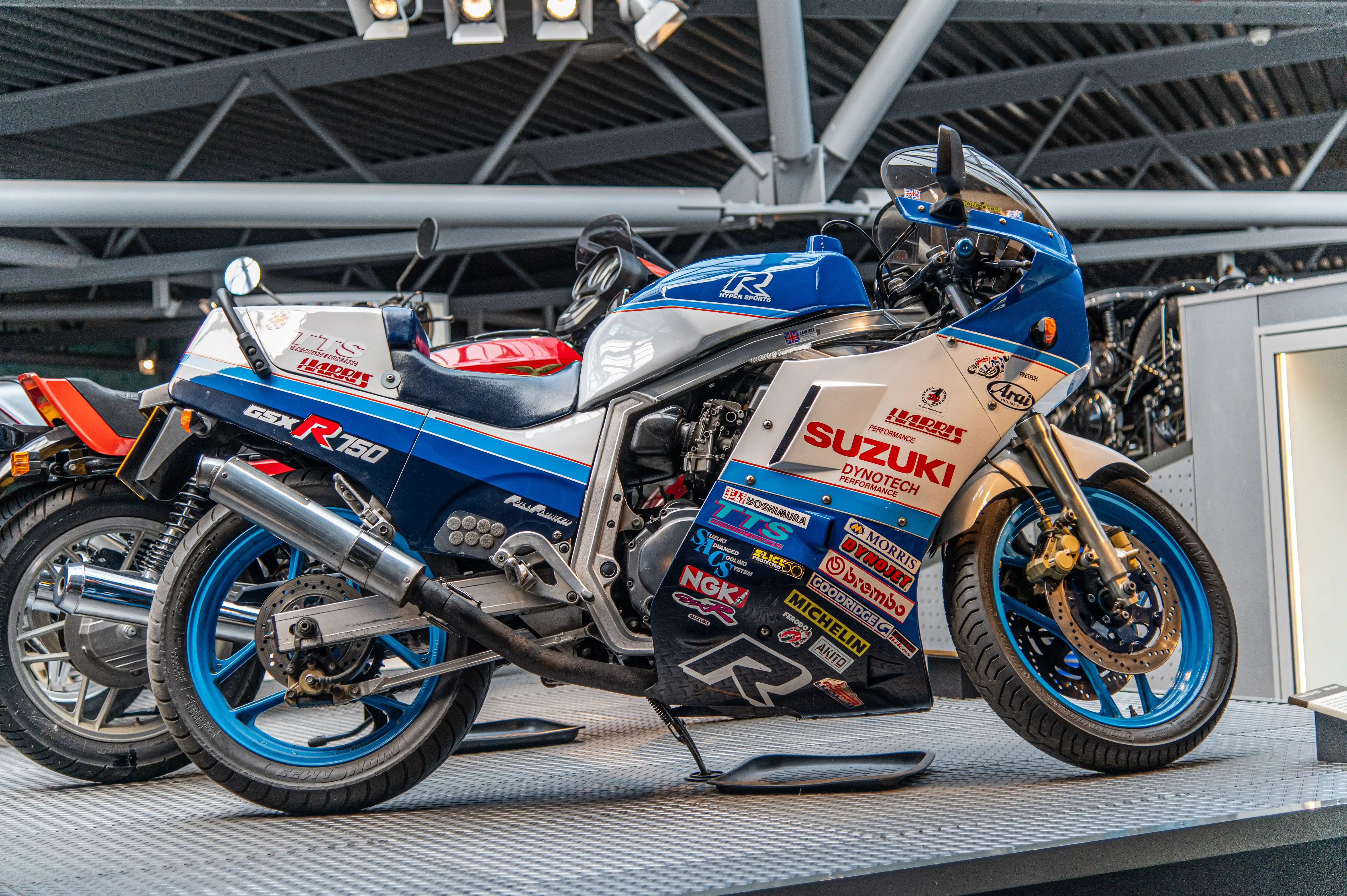 The image showcases a Suzuki GSX-R750 motorcycle, prominently displayed in a well-lit exhibition space. The motorcycle features a distinctive blue and white color scheme with numerous sponsor decals, including brands like Harris Performance, Dynotech, and Michelin. The bike is equipped with a single seat and various performance enhancements, indicating it is likely a racing or high-performance model. The setting appears to be an indoor venue, possibly a motorcycle show or museum, with other motorcycles visible in the background.