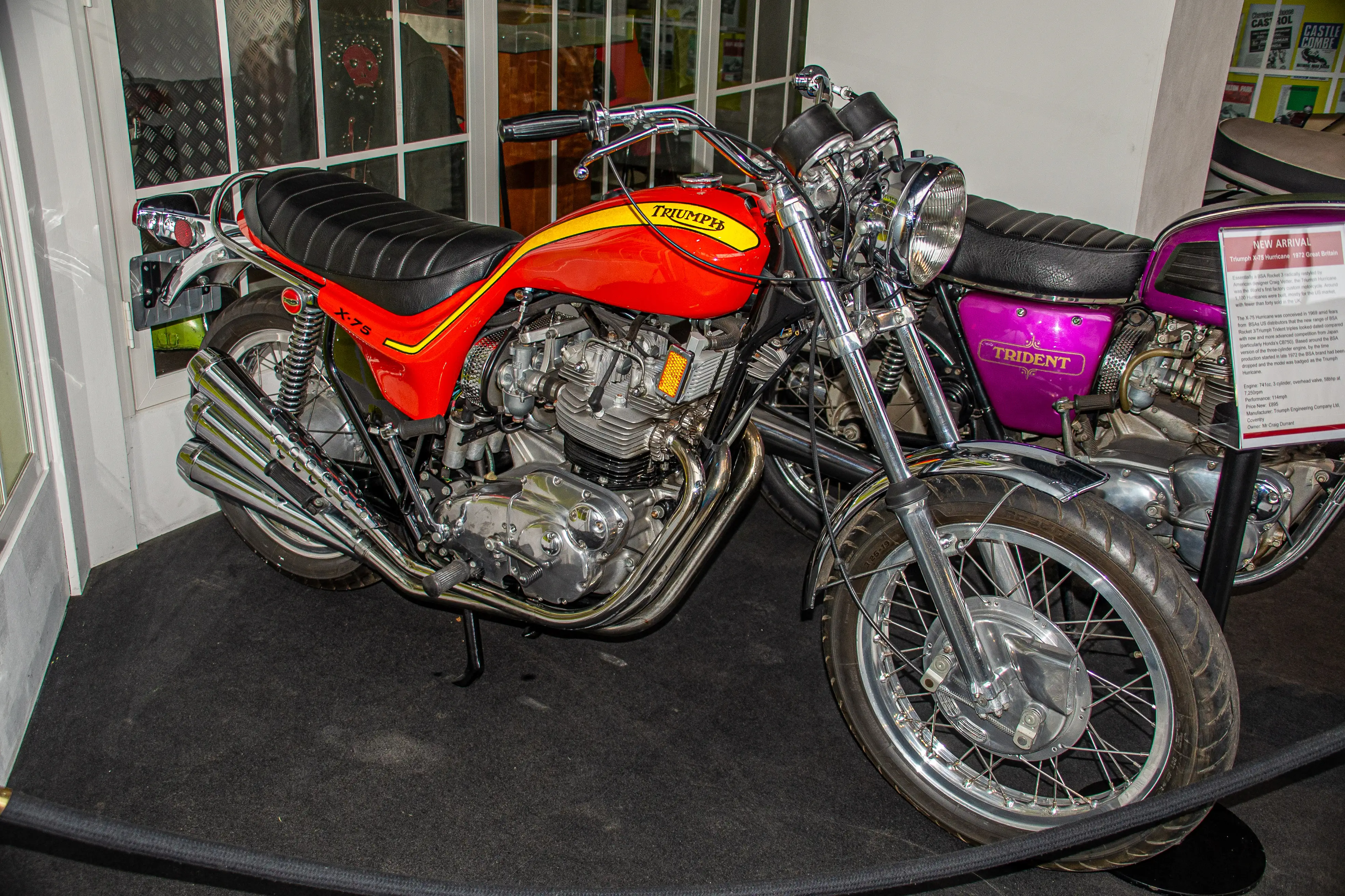 The image shows two vintage Triumph motorcycles on display. The motorcycle in the foreground is a Triumph Trident, characterized by its distinctive purple and chrome color scheme. The motorcycle behind it is a Triumph Bonneville, easily identifiable by its red and yellow paint job. Both motorcycles are displayed indoors, likely in a museum or showroom, with detailed information placards next to them. The motorcycles feature classic designs with polished chrome engines and black leather seats.