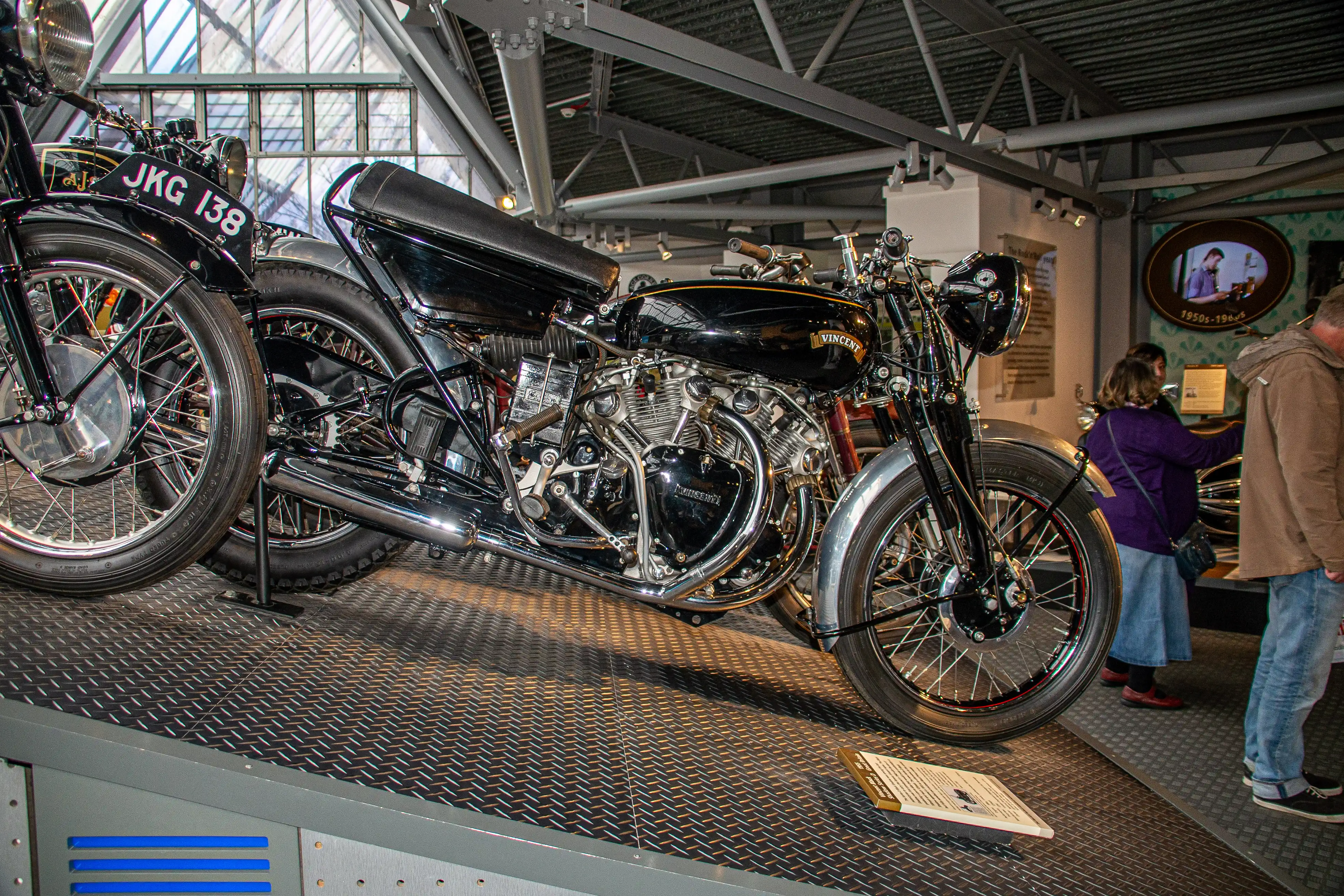 The image depicts a vintage motorcycle exhibit in a museum. The motorcycle is a Vincent, a classic model from the mid-20th century, known for its distinctive design and engineering. The exhibit is housed in a well-lit, modern space with a glass ceiling, allowing natural light to illuminate the motorcycle. Two individuals are observing the motorcycle, reading the information provided on placards. The setting suggests an educational environment focused on showcasing historical motorcycles.
