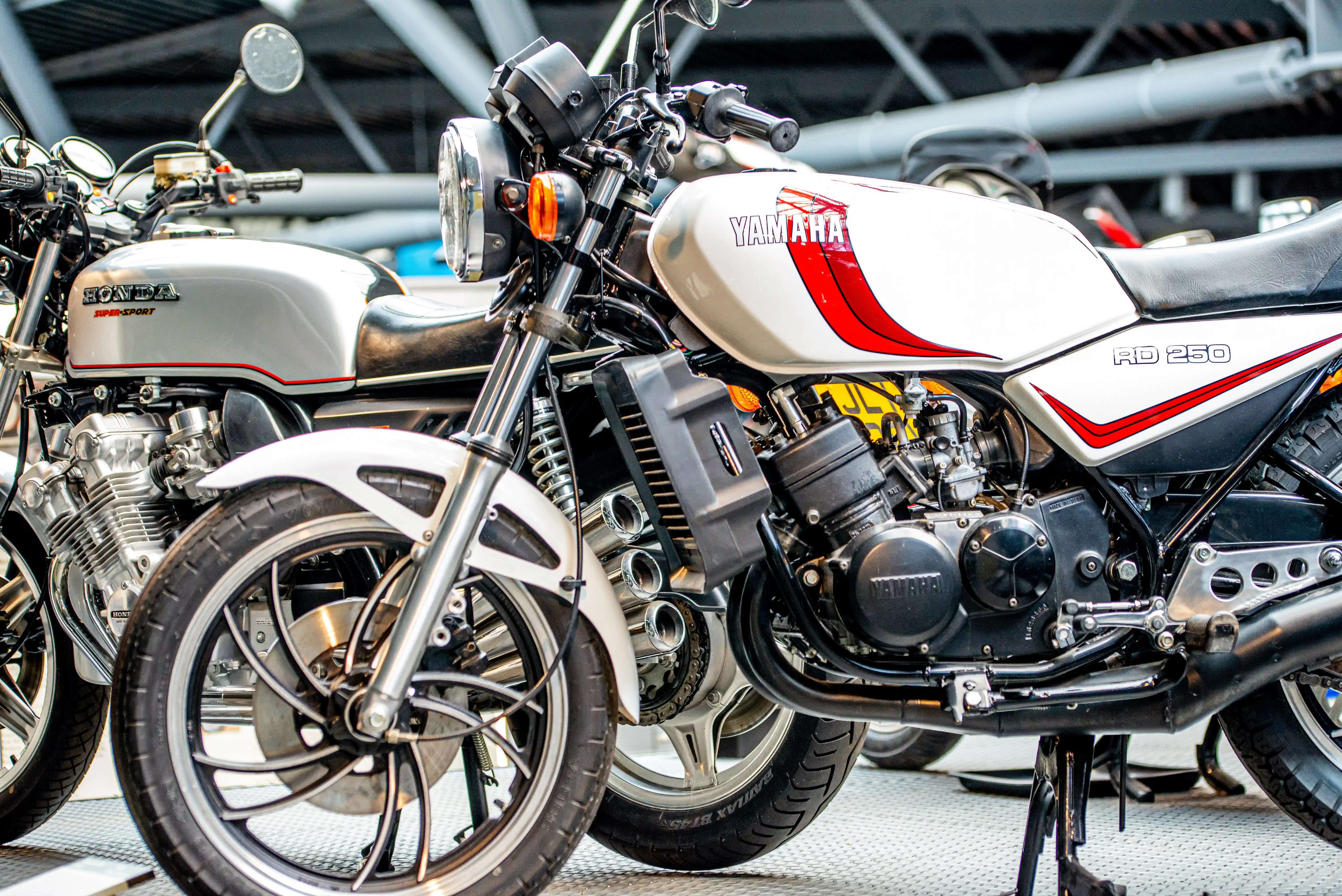 The image shows two motorcycles parked side by side. The motorcycle on the left is a Honda with the model name 'Sport' visible on its body. The motorcycle on the right is a Yamaha with the model name 'RD 250' visible on its body. Both motorcycles are displayed in a well-lit indoor setting, likely an exhibition or showroom.