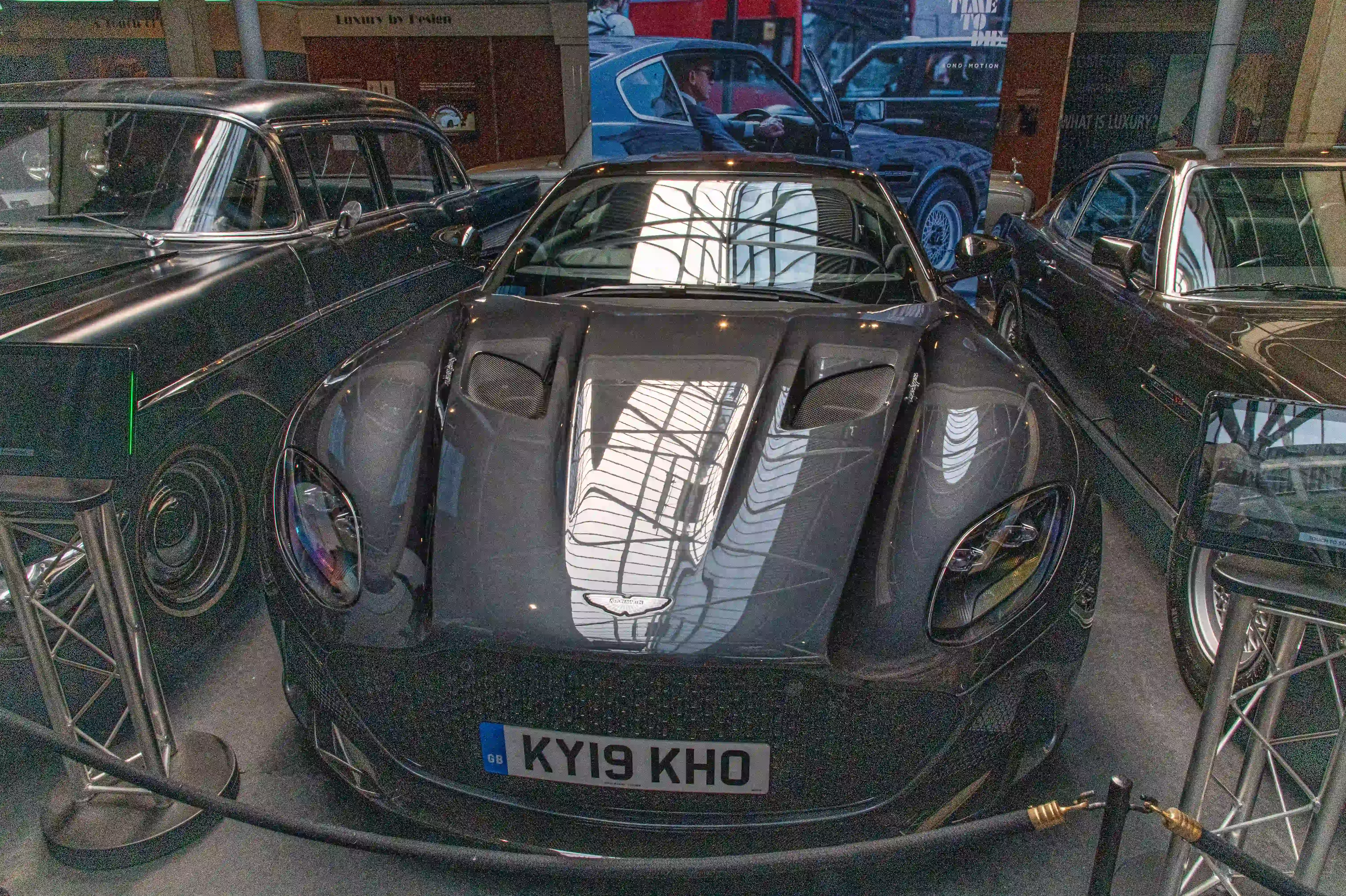 The image shows a sleek, black sports car displayed in a museum or exhibition setting. The car has a distinctive, aerodynamic design with prominent air vents on the hood and a glossy finish. The license plate reads 'KYI9 KHO.' The car is surrounded by a protective barrier, indicating it is a valuable or significant exhibit. The background includes other classic cars and exhibition banners, suggesting a theme of luxury and design.