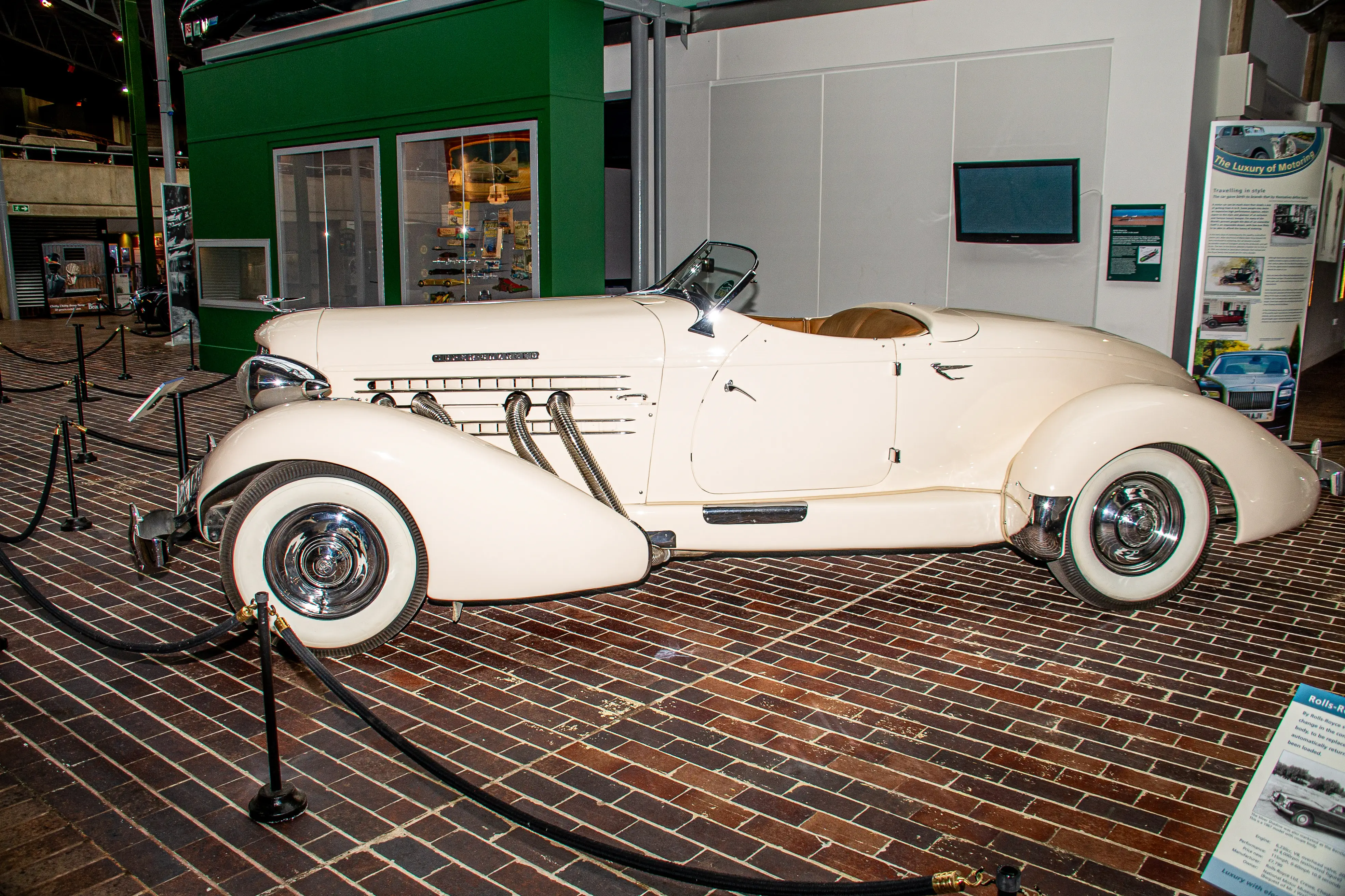 The image depicts a classic, cream-colored convertible car on display in a museum setting. The car features prominent, exposed exhaust pipes, a sleek, vintage design, and is cordoned off with black ropes and stanchions. The surrounding area includes informational displays and a glimpse into an adjacent room filled with various other vehicles and exhibits.