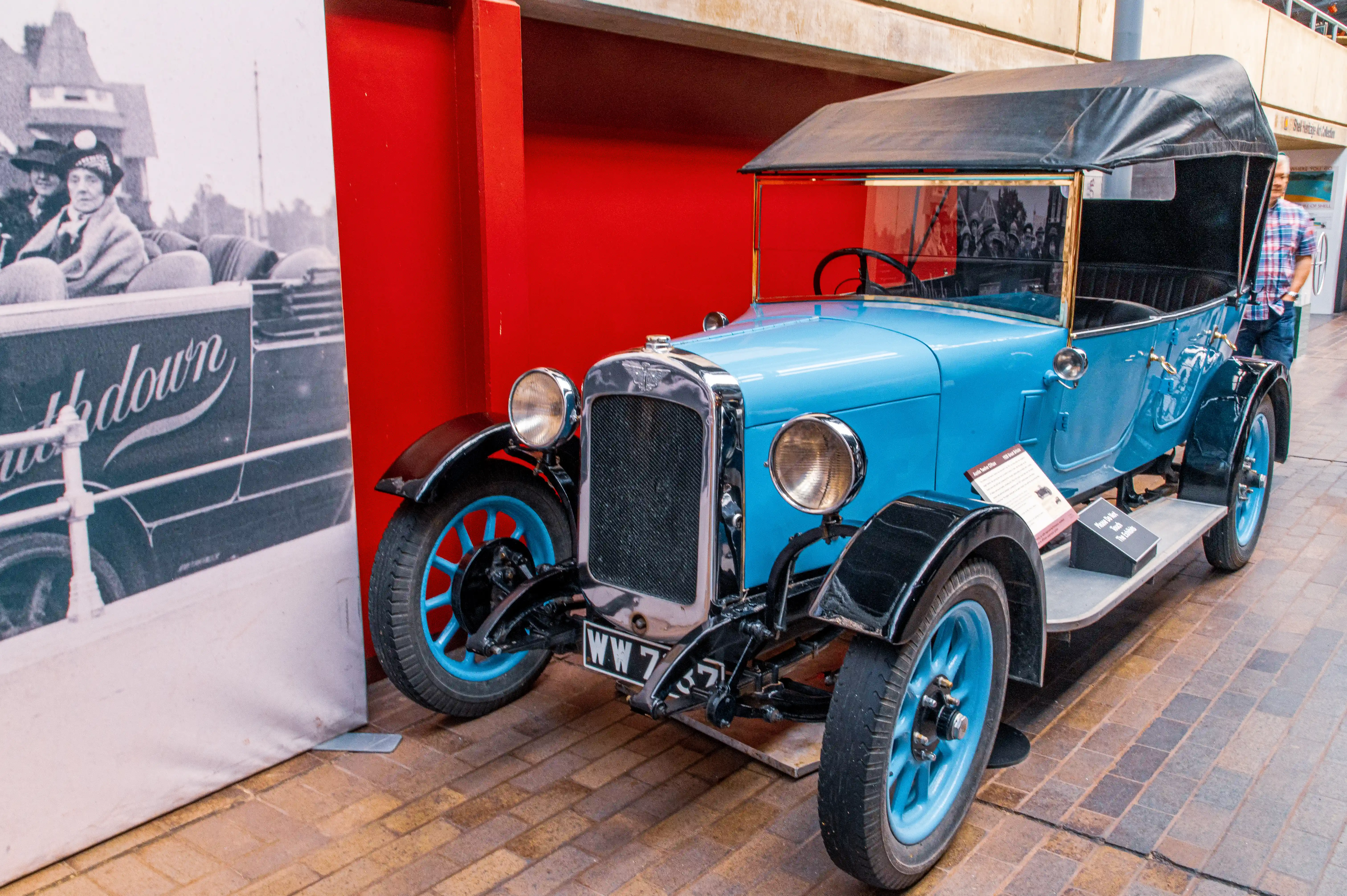 The image shows a vintage blue car on display, likely at a museum or exhibition. The car has a classic design with a black convertible top, large round headlights, and wire-spoke wheels. It is parked on a brick pavement in front of a red wall and a large black-and-white photograph. The photograph depicts people from an earlier era, suggesting a historical theme. The car's license plate reads 'WW 7'.