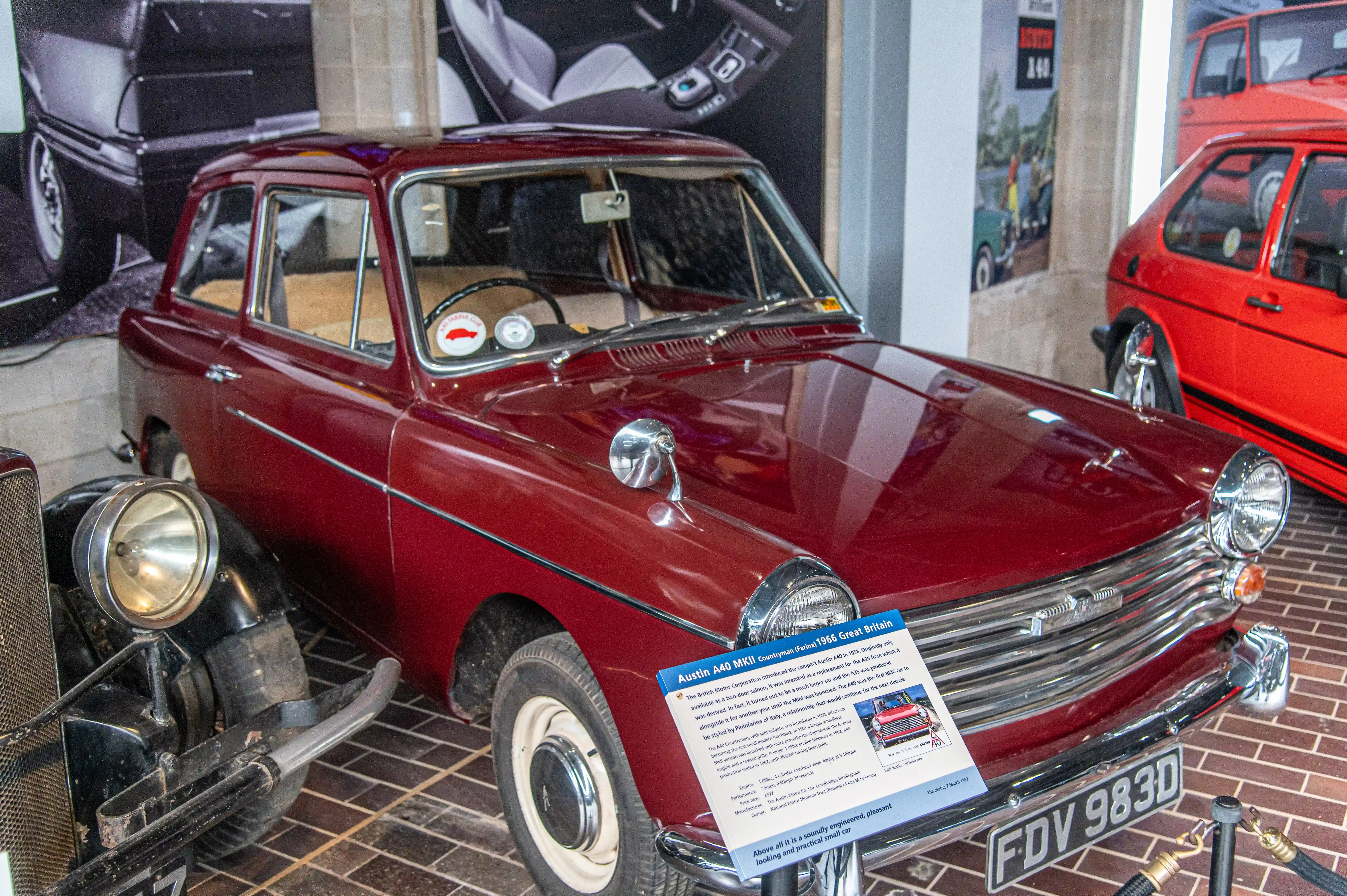 The image shows a classic Austin A40 MKII car on display in a museum. The car is maroon in color and has a sign in front of it providing information about its history and features. The sign indicates that the Austin A40 MKII was introduced by the British Motor Corporation in 1961 as a compact and economical vehicle. It highlights the car's practicality, affordability, and its significance in automotive history.
